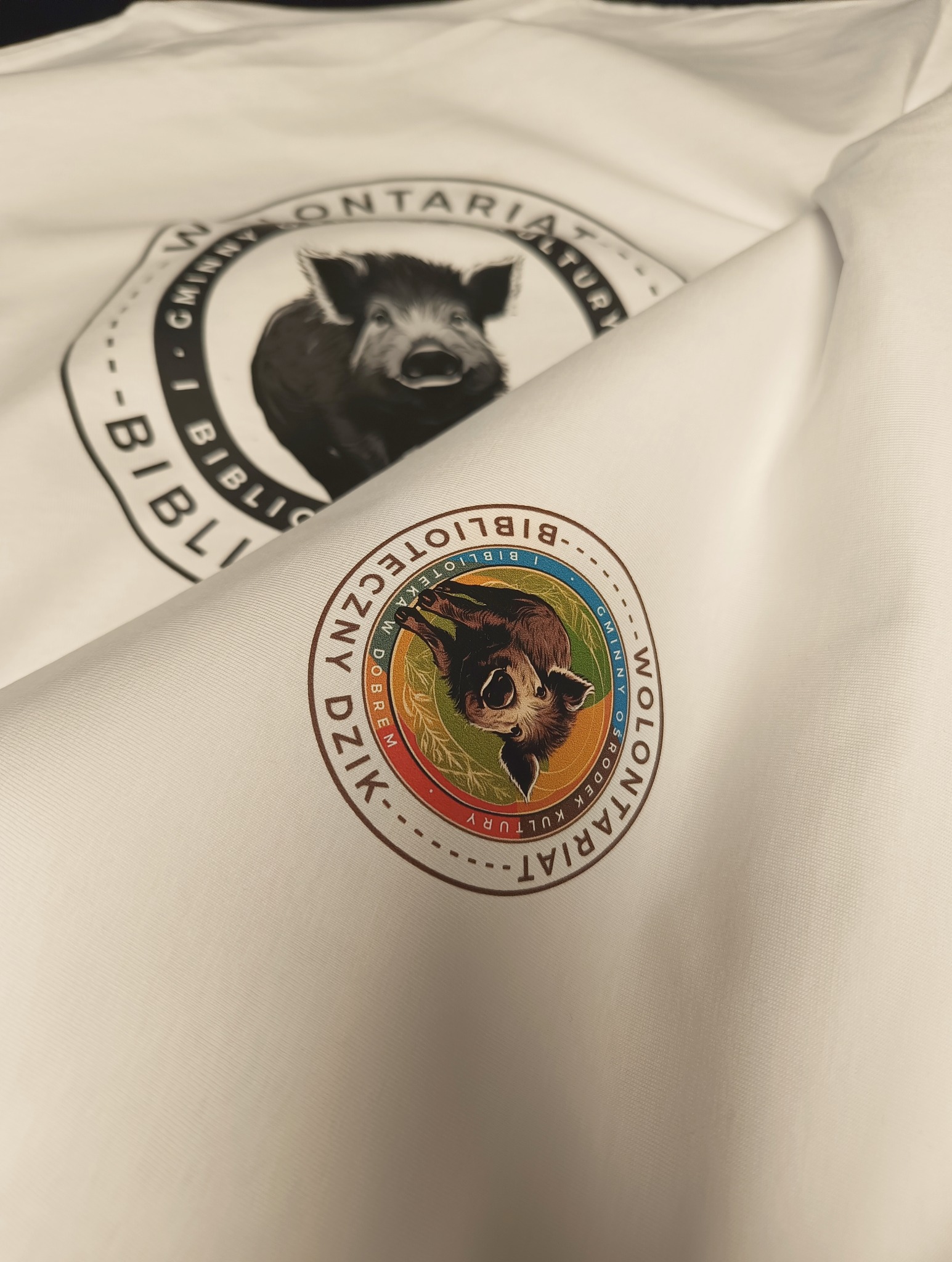 Biała koszulka z nadrukiem logotypu 'Biblioteka i Gminny Ośrodek Kultury' z motywem dzika, widoczne dwa różne warianty logotypu.