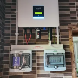 Biały inwerter FOX z wyświetlaczem i logo NEX ENERGY zamontowany na ścianie z brązowych płytek, poniżej dwie rozdzielnice PV-AC i PV-DC marki Noark z widocznymi zabezpieczeniami.