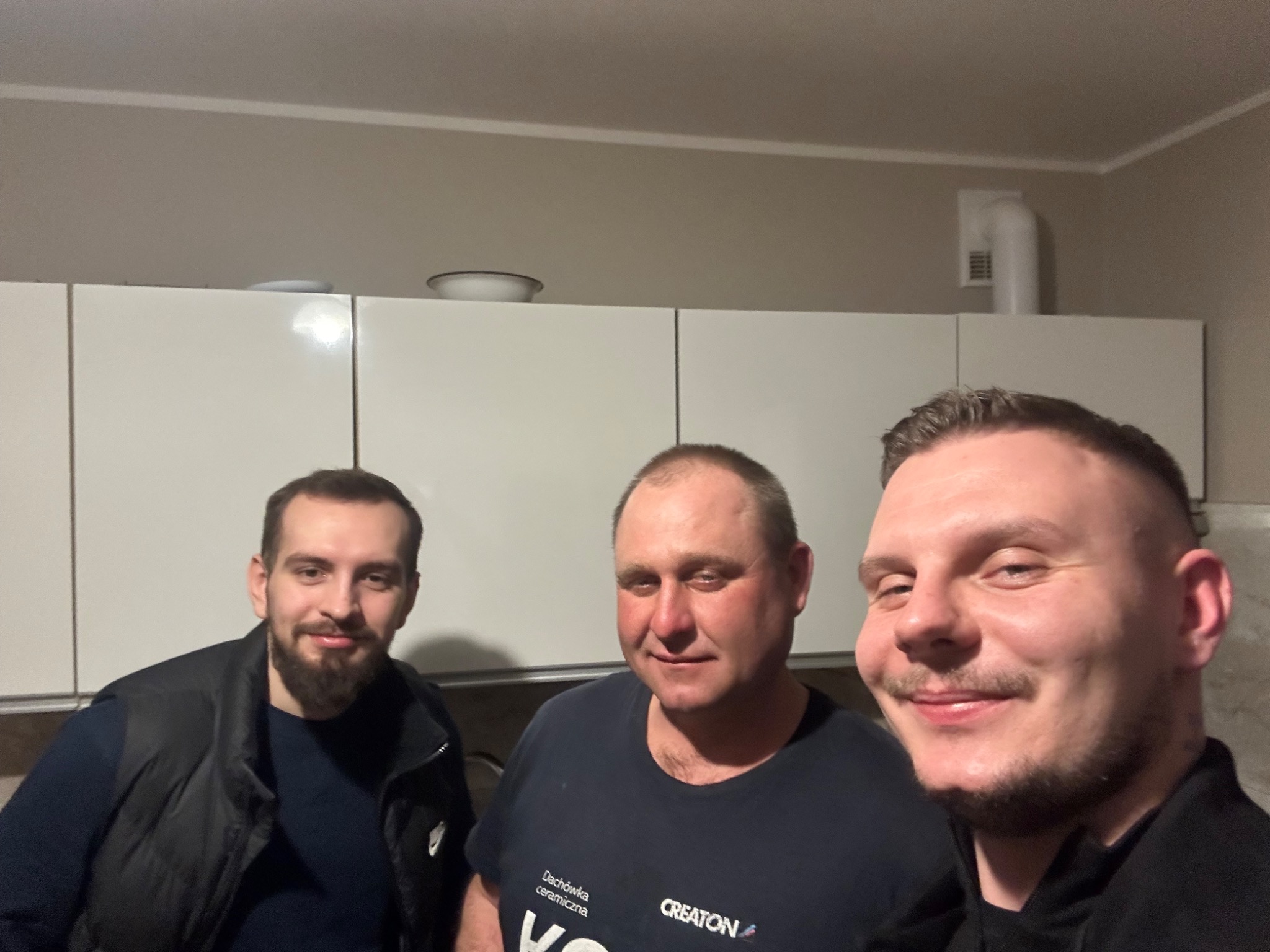 Trzech mężczyzn uśmiechniętych do kamery, w tle białe szafki kuchenne, jeden z nich ma na sobie koszulkę firmy CREATON Dachówka ceramiczna.