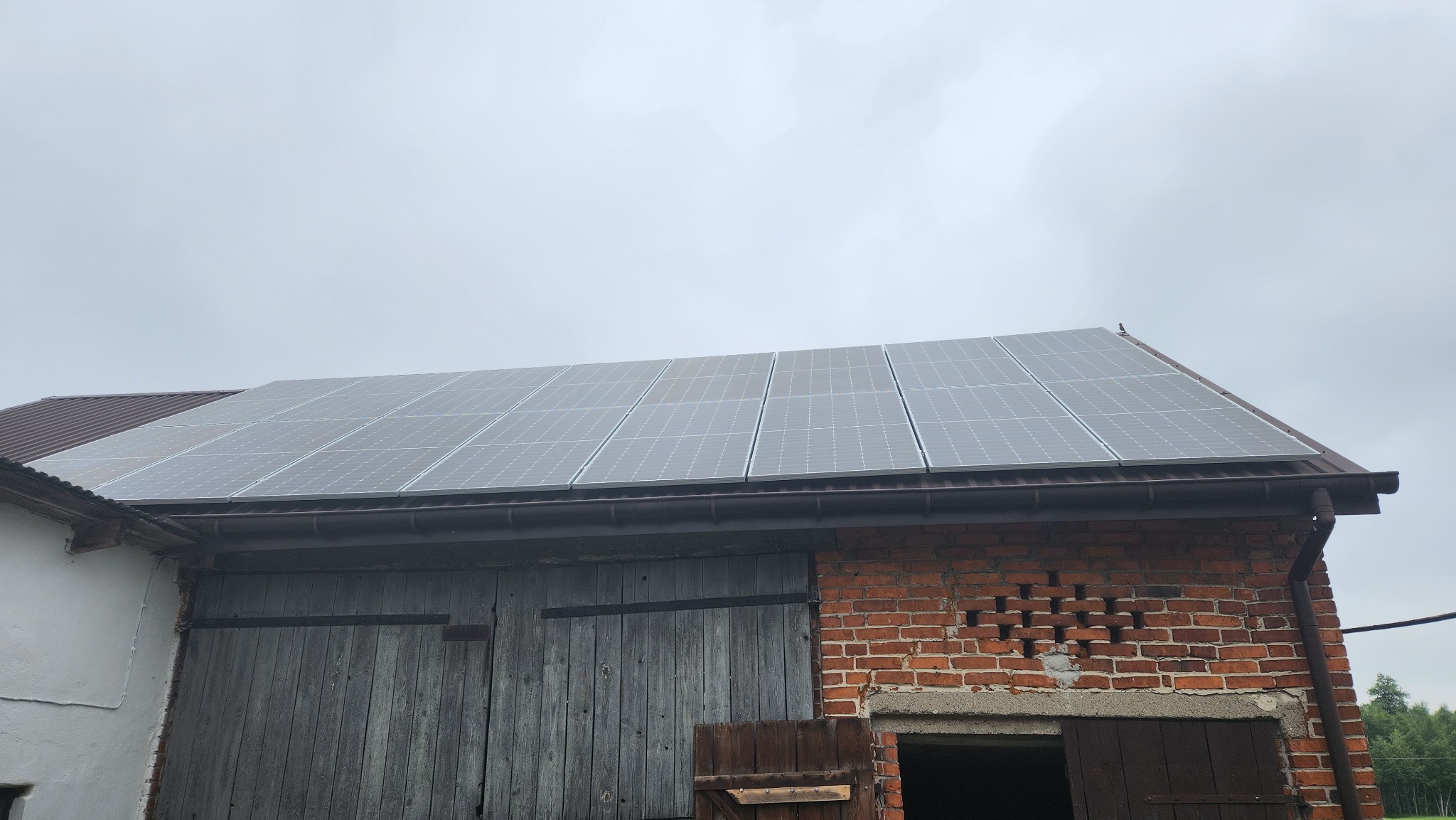 Instalacja paneli słonecznych na dachu starej stodoły z cegły i drewna, widok z dołu na pochmurnym niebie.