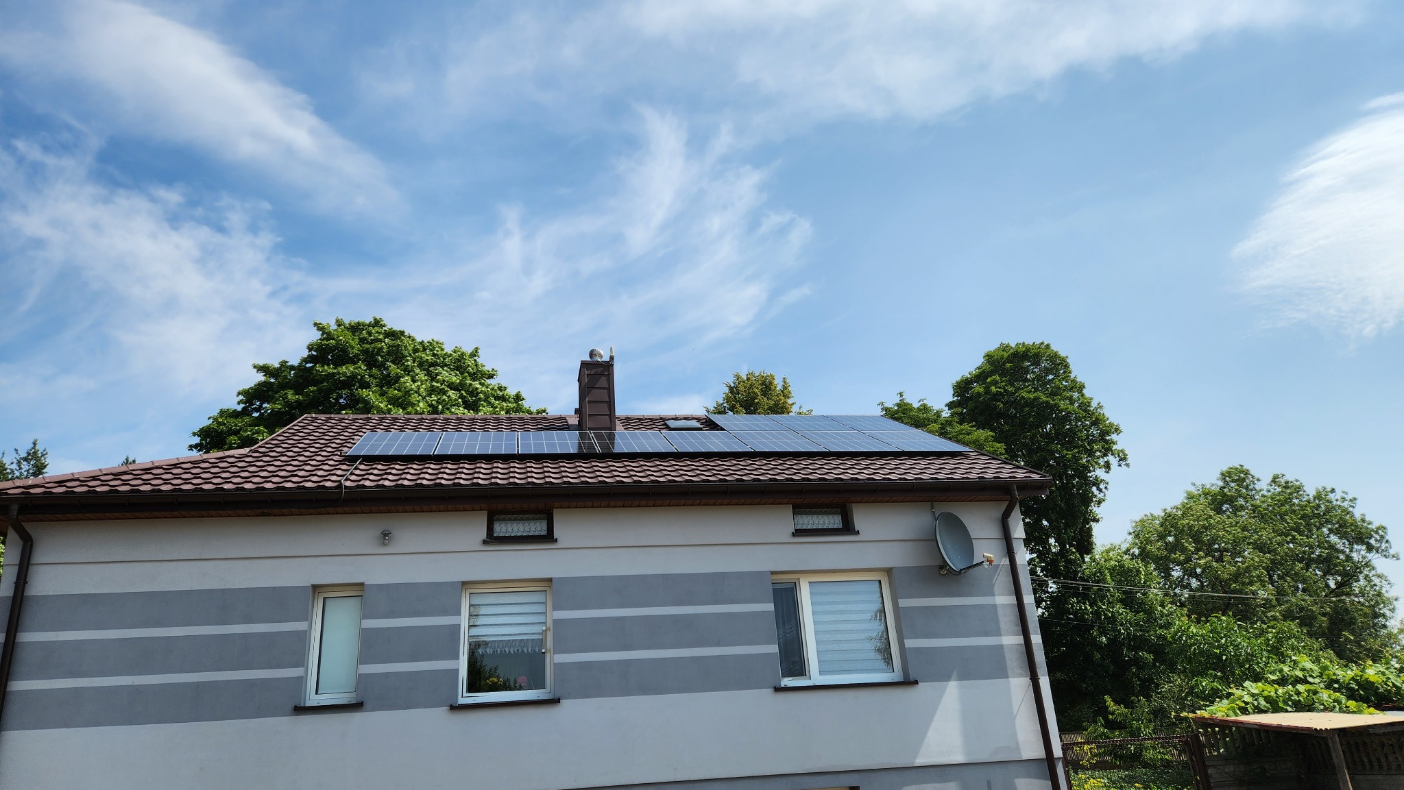 Panele słoneczne na brązowym dachu dwupiętrowego domu z szarymi pasami, widoczne z poziomu gruntu pod błękitnym niebem z delikatnymi chmurami.