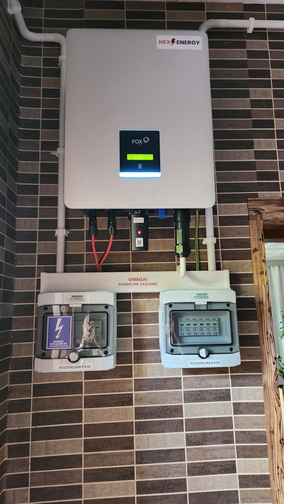 Biały inwerter FOX z wyświetlaczem i logo NEX ENERGY zamontowany na ścianie z brązowych płytek, poniżej dwie rozdzielnice PV-AC i PV-DC marki Noark z widocznymi zabezpieczeniami.
