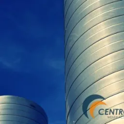 Dwa srebrne, cylindryczne zbiorniki z poziomymi żłobieniami na tle intensywnie błękitnego nieba z delikatnymi smugami chmur. Na jednym ze zbiornik&oacute;w widoczne logo firmy 'CENTROWENT WENTYLACJA...