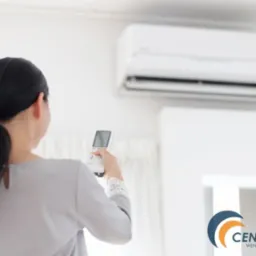 Kobieta z tyłu, trzymająca pilot klimatyzacji, ustawia temperaturę w pomieszczeniu. W tle biały klimatyzator ścienny i białe ściany. W prawym dolnym rogu widoczne logo firmy.