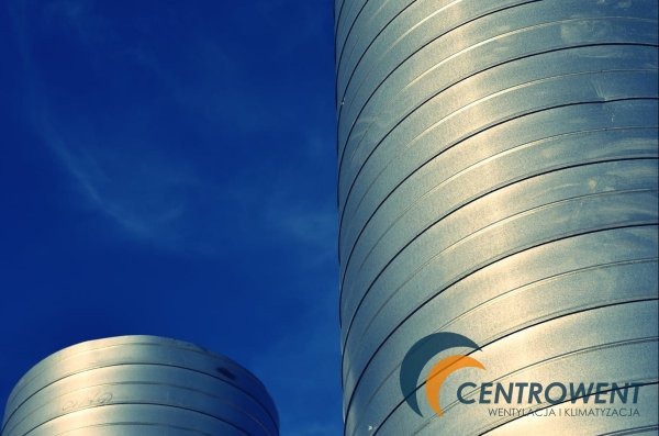 Dwa srebrne, cylindryczne zbiorniki z poziomymi żłobieniami na tle intensywnie błękitnego nieba z delikatnymi smugami chmur. Na jednym ze zbiorników widoczne logo firmy 'CENTROWENT WENTYLACJA...