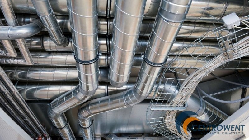 Srebrne, spiralne rury wentylacyjne pod sufitem, z widocznymi kablami w metalowej siatce i logo firmy CENTROWENT w prawym dolnym rogu.
