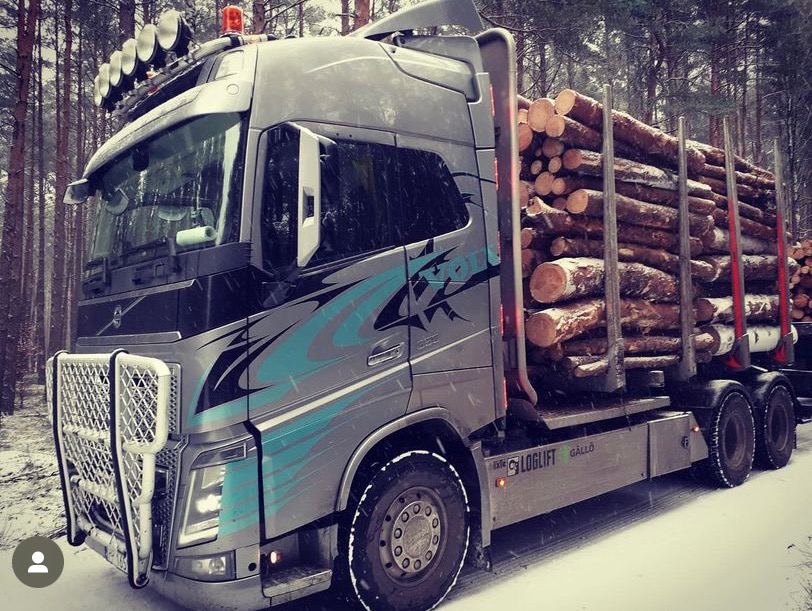Ciężarówka Volvo z niebieskimi grafikami na boku, załadowana drewnem, stoi na ośnieżonej drodze w lesie.