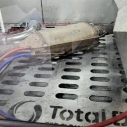 Filtr cząstek stałych (DPF) w komorze czyszczącej z widocznymi przewodami i logo Total DPF Clean na perforowanej podstawie.