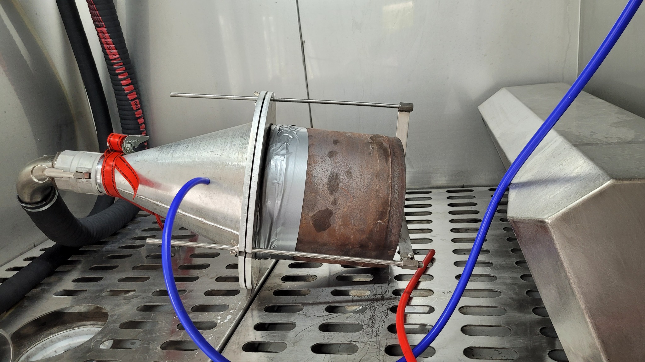 Filtr cząstek stałych (DPF) zabezpieczony taśmą, zamocowany w urządzeniu do czyszczenia, z widocznymi przewodami doprowadzającymi płyny.