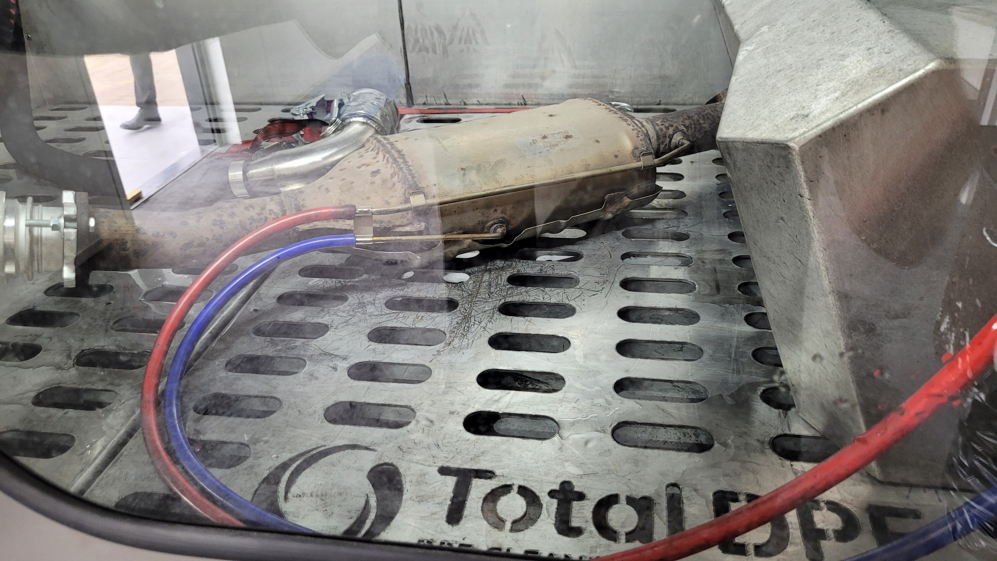 Filtr cząstek stałych (DPF) w komorze czyszczącej z widocznymi przewodami i logo Total DPF Clean na perforowanej podstawie.