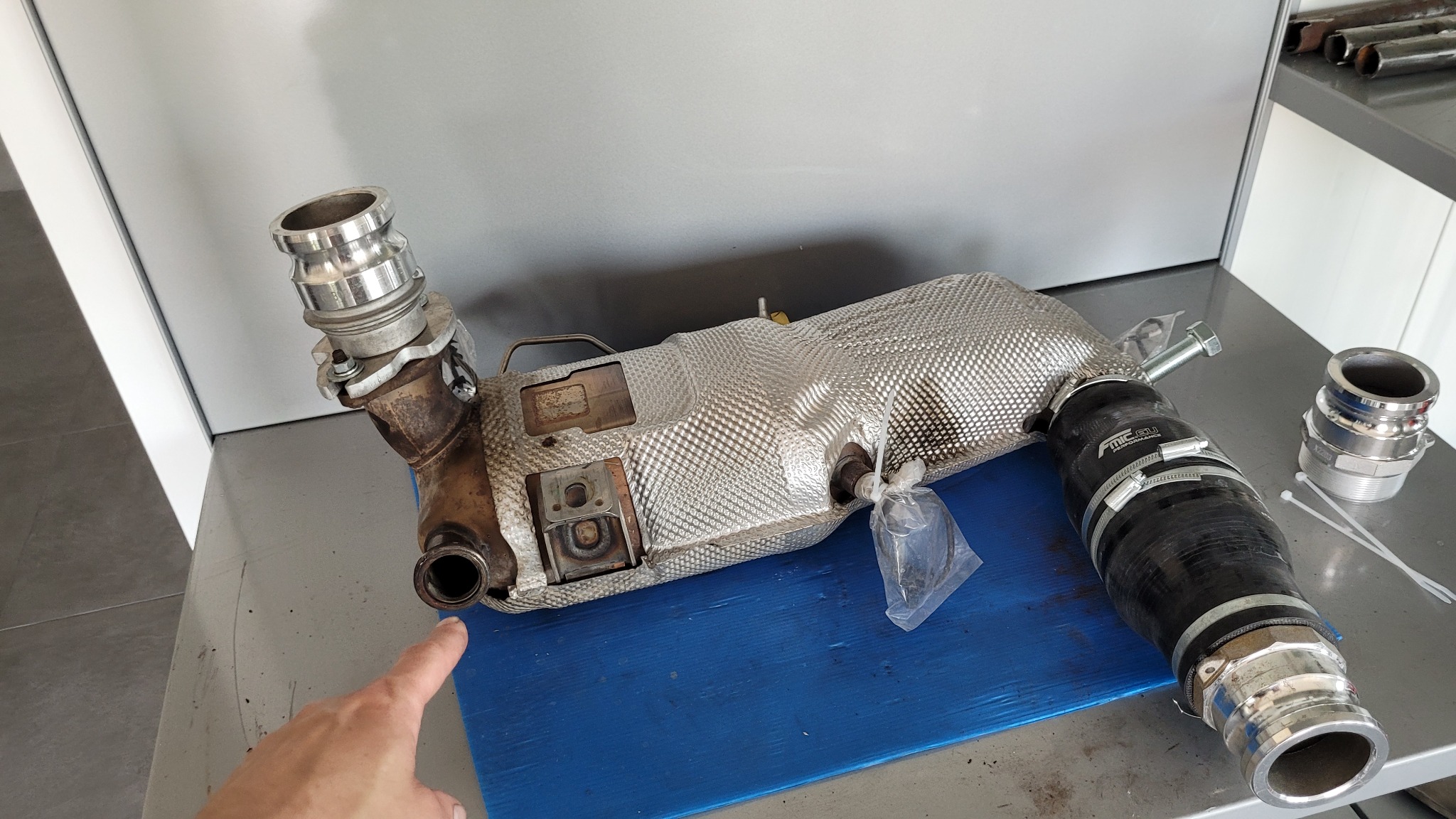 Wymontowany katalizator samochodowy z elementami montażowymi na stole warsztatowym, wskazany palcem.