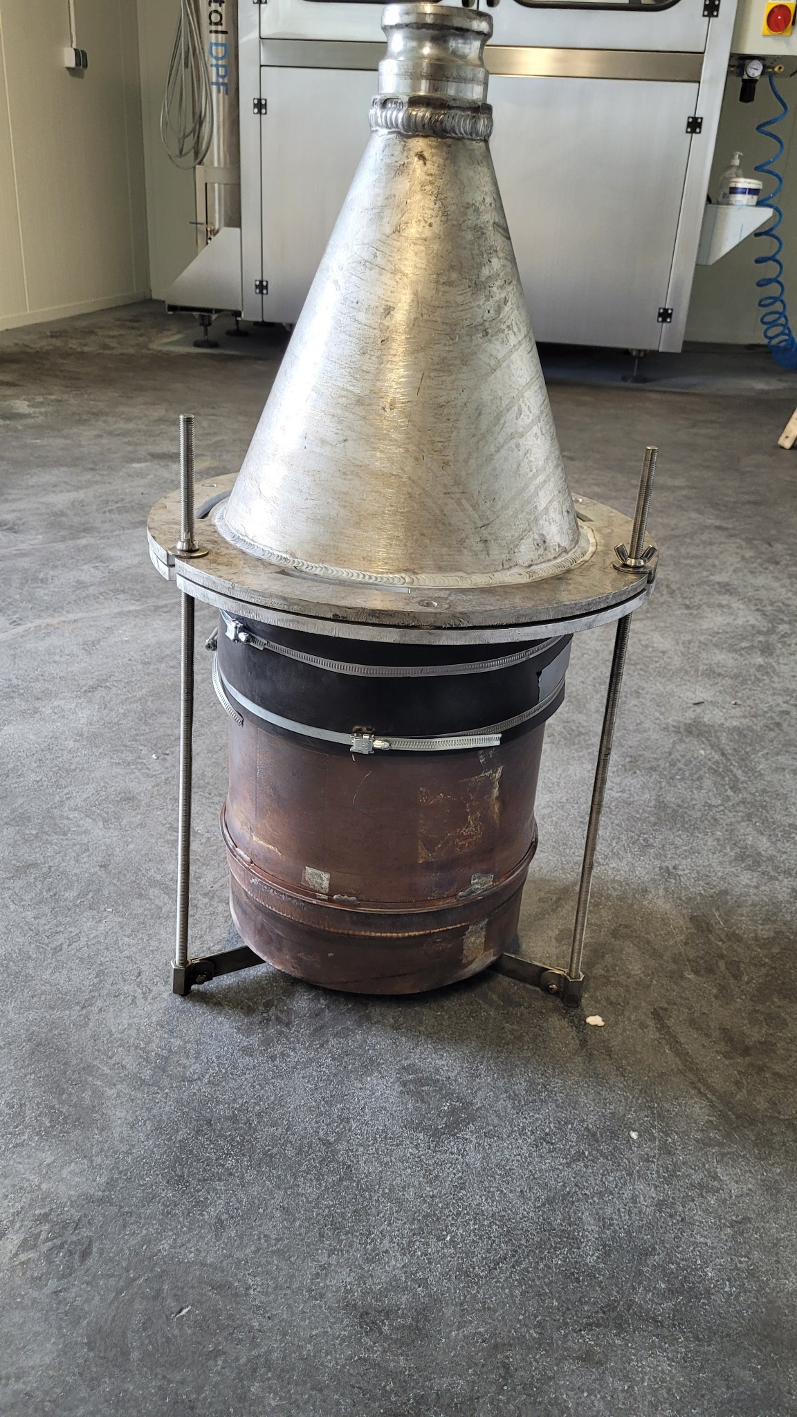 Metalowa konstrukcja z elementami spawanymi, składająca się z stożka i cylindra, na metalowym stelażu, widoczne ślady użytkowania i spawania, w tle urządzenia przemysłowe.