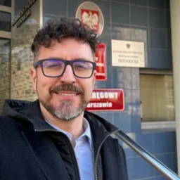 Uśmiechnięty mężczyzna w okularach pozuje przed budynkiem Kancelarii Notarialnej z godłem Polski w tle, widoczny fragment tablicy informacyjnej oraz metalowa poręcz.