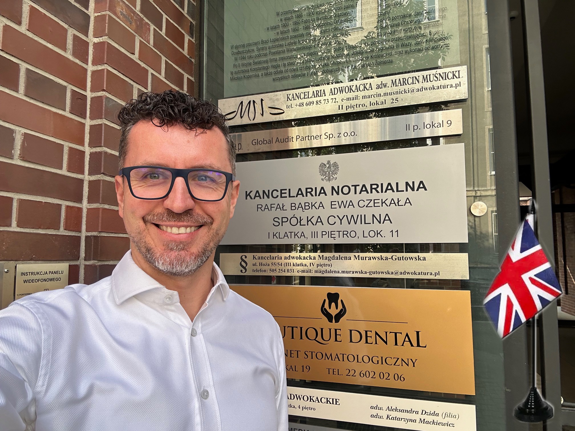 Uśmiechnięty mężczyzna w okularach pozuje na tle tablic informacyjnych kancelarii prawnych i notarialnych, z widoczną flagą Wielkiej Brytanii.