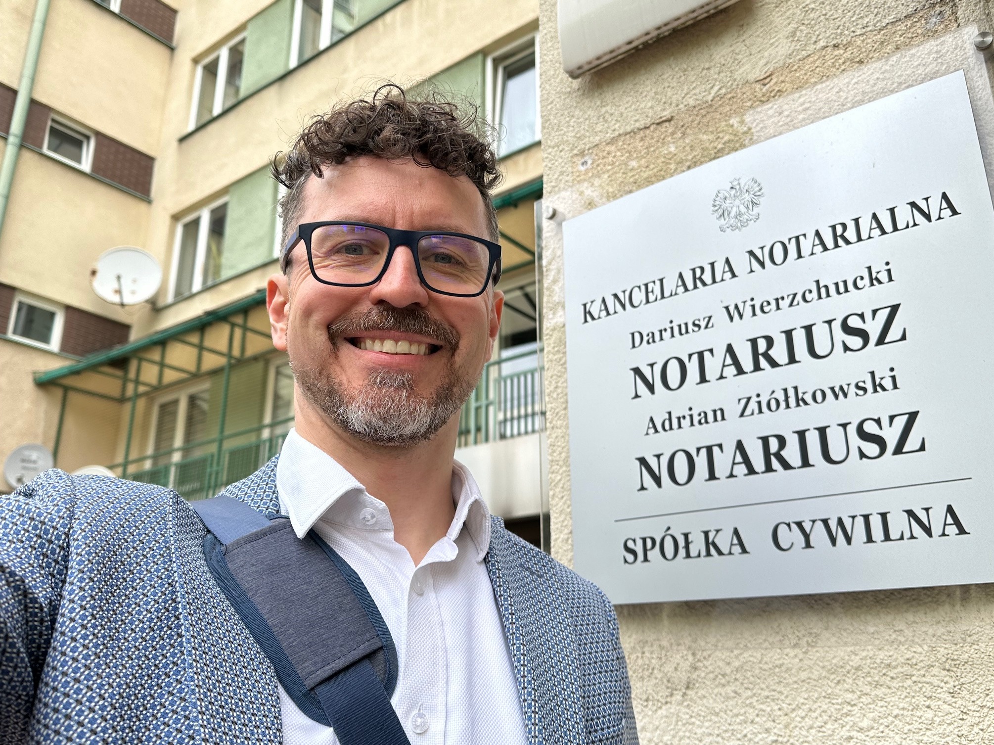 Uśmiechnięty mężczyzna w okularach i marynarce stoi przed tablicą informacyjną Kancelarii Notarialnej Dariusza Wierzchuckiego i Adriana Ziółkowskiego.