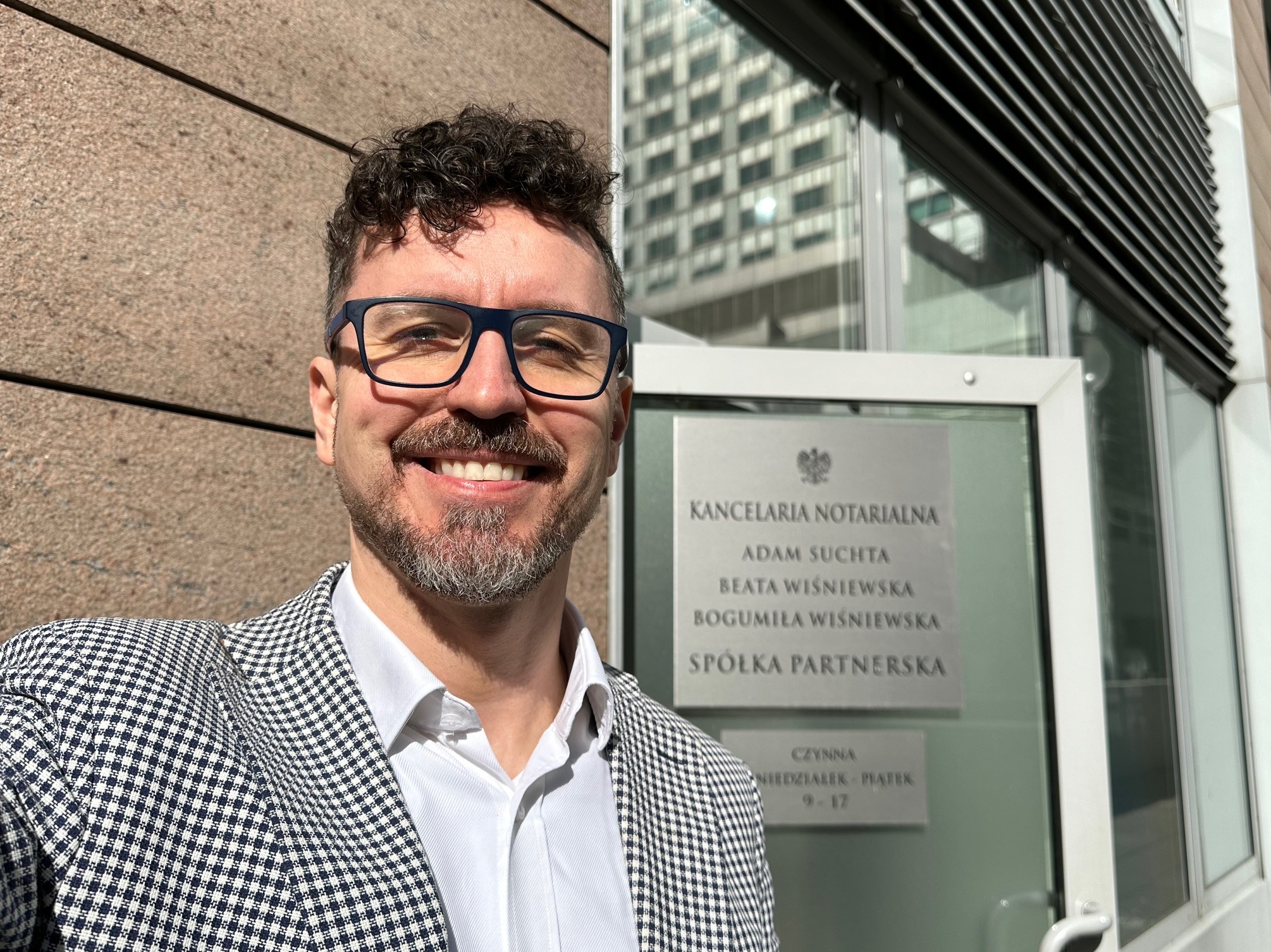 Uśmiechnięty mężczyzna w okularach i marynarce w kratę stoi przed wejściem do kancelarii notarialnej z tabliczką informacyjną z nazwiskami notariuszy i godzinami otwarcia.