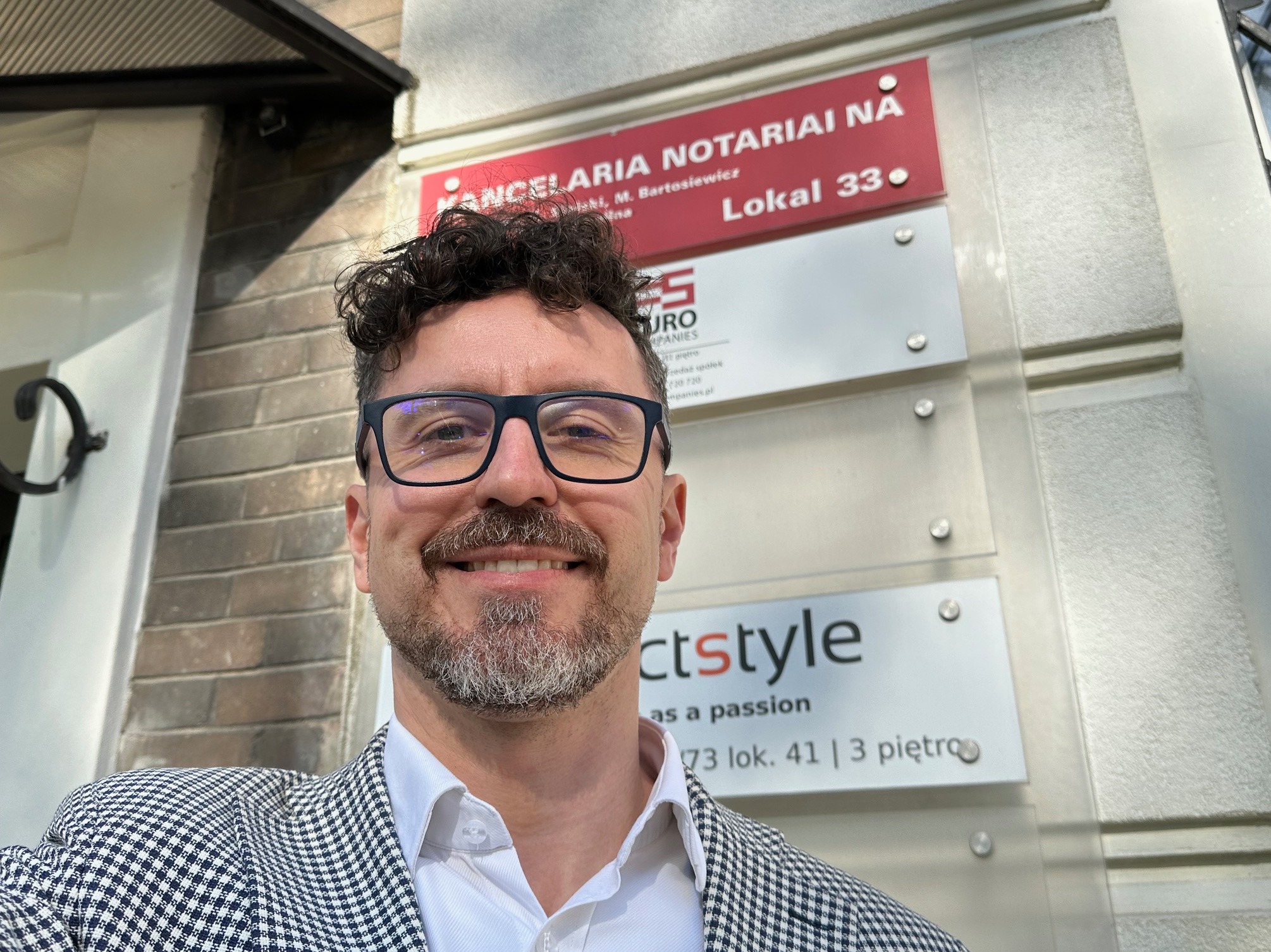 Mężczyzna w okularach i marynarce w kratę robi selfie na tle tablic informacyjnych firm, w tym kancelarii notarialnej Lokal 33 i firmy o nazwie 'ctstyle'.