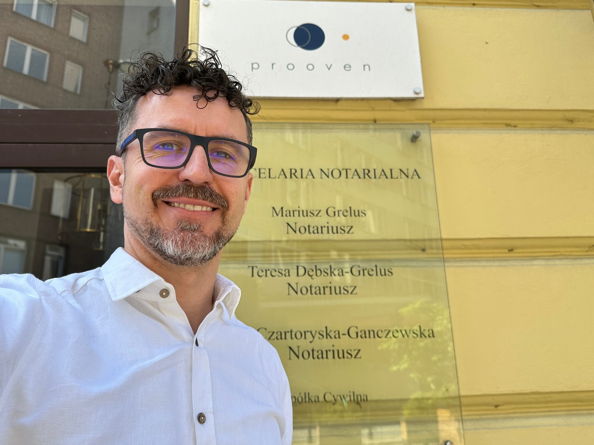 Uśmiechnięty mężczyzna w okularach i białej koszuli pozuje przed budynkiem z tablicą informacyjną Kancelarii Notarialnej z nazwiskami notariuszy: Mariusz Grelus, Teresa Dębska-Grelus...