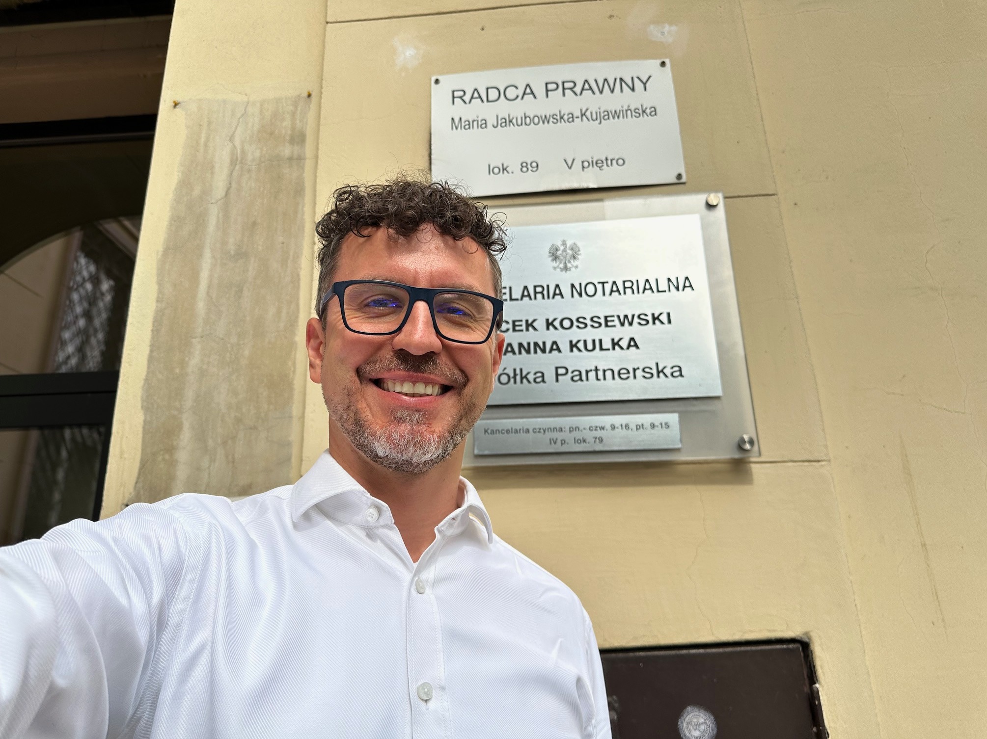 Uśmiechnięty mężczyzna w okularach i białej koszuli robi selfie na tle tabliczek informacyjnych kancelarii radcy prawnego i notarialnej w budynku.