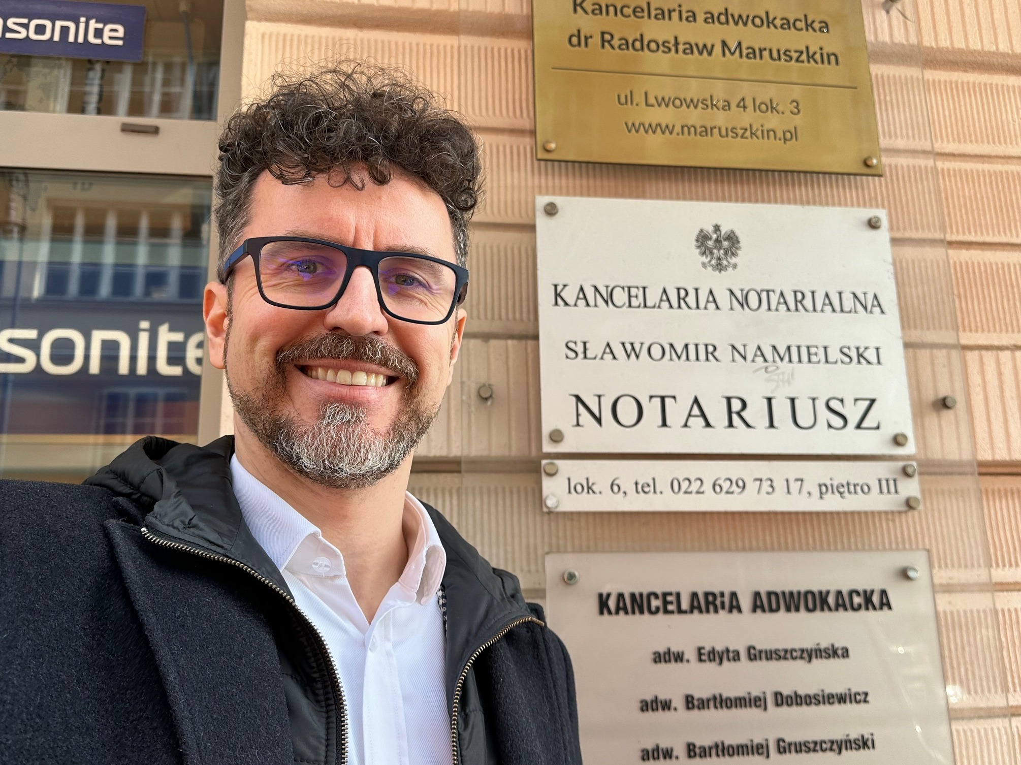 Uśmiechnięty mężczyzna w okularach robi selfie na tle tablic informacyjnych kancelarii adwokackiej i notarialnej na ulicy Lwowskiej.