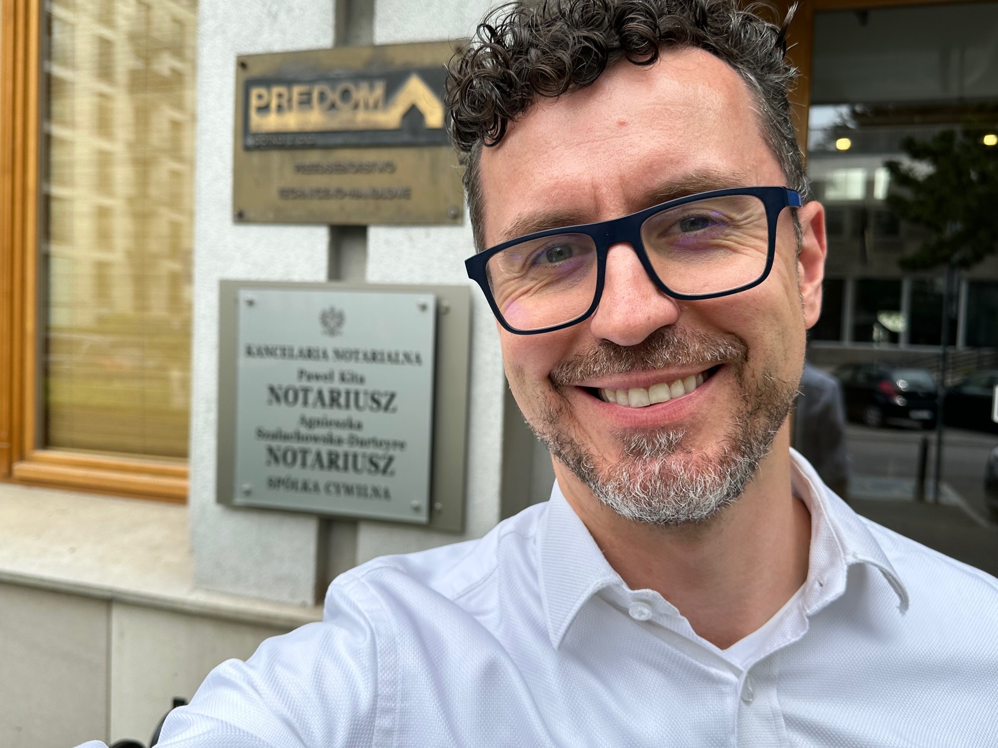 Uśmiechnięty mężczyzna w okularach na tle budynku Kancelarii Notarialnej w Warszawie.