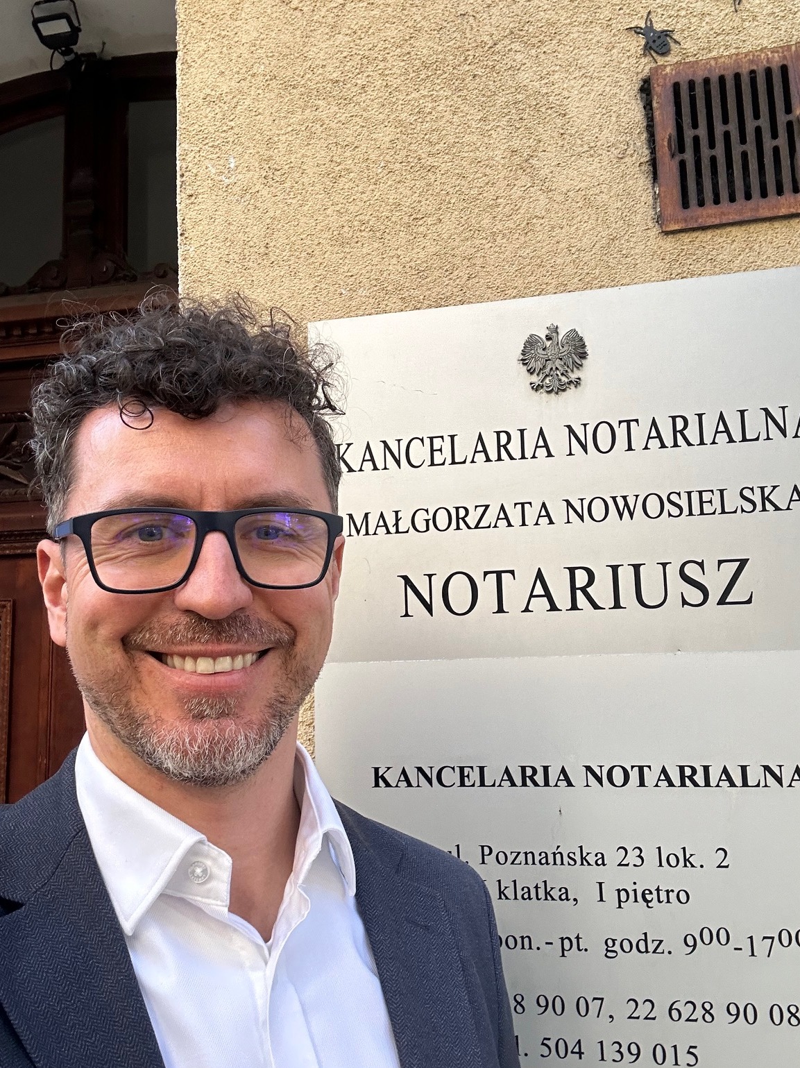 Uśmiechnięty mężczyzna w okularach i marynarce pozuje na tle tablicy informacyjnej Kancelarii Notarialnej Małgorzaty Nowosielskiej z adresem i godzinami otwarcia.