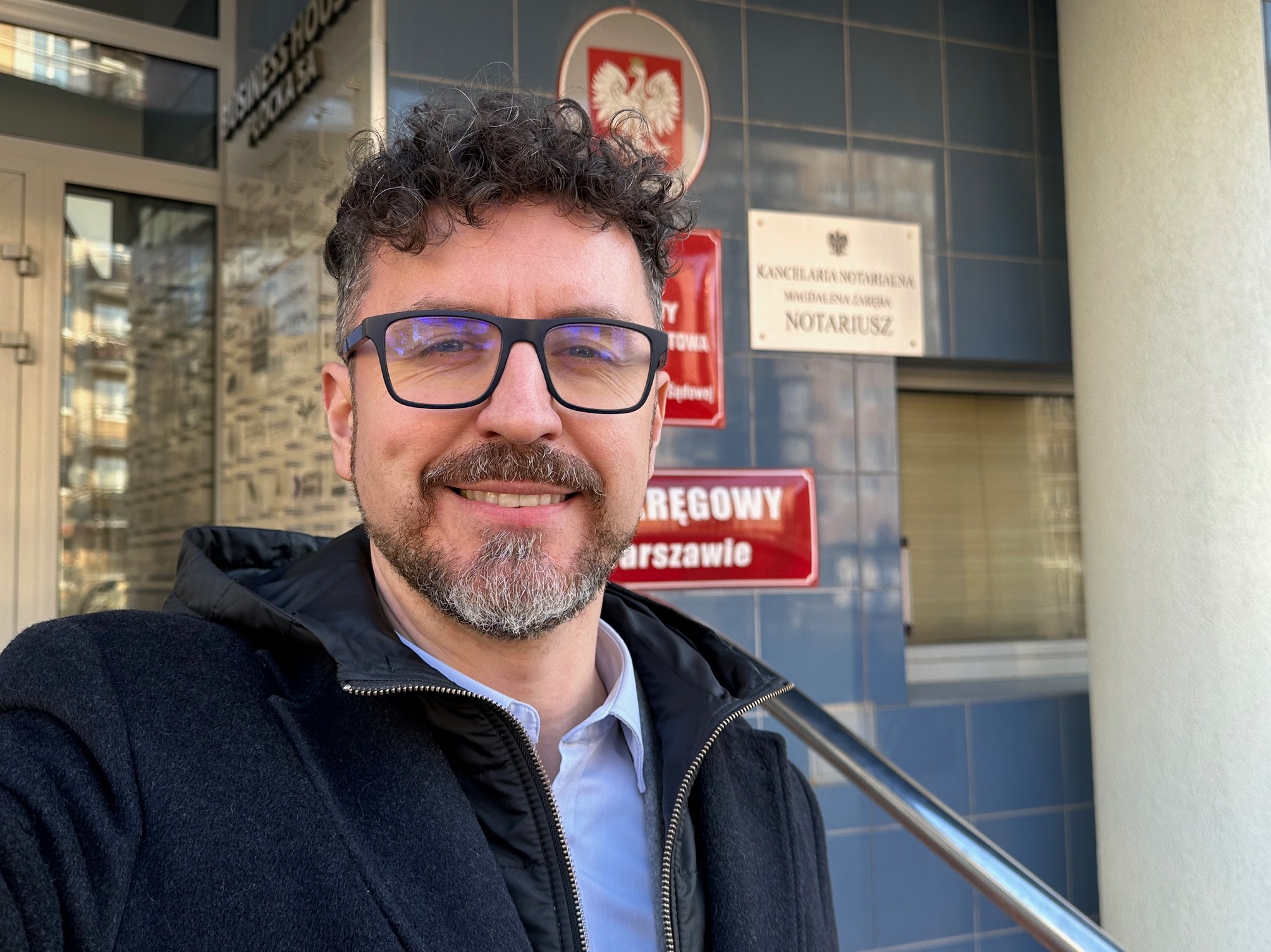 Uśmiechnięty mężczyzna w okularach pozuje przed budynkiem Kancelarii Notarialnej z godłem Polski w tle, widoczny fragment tablicy informacyjnej oraz metalowa poręcz.