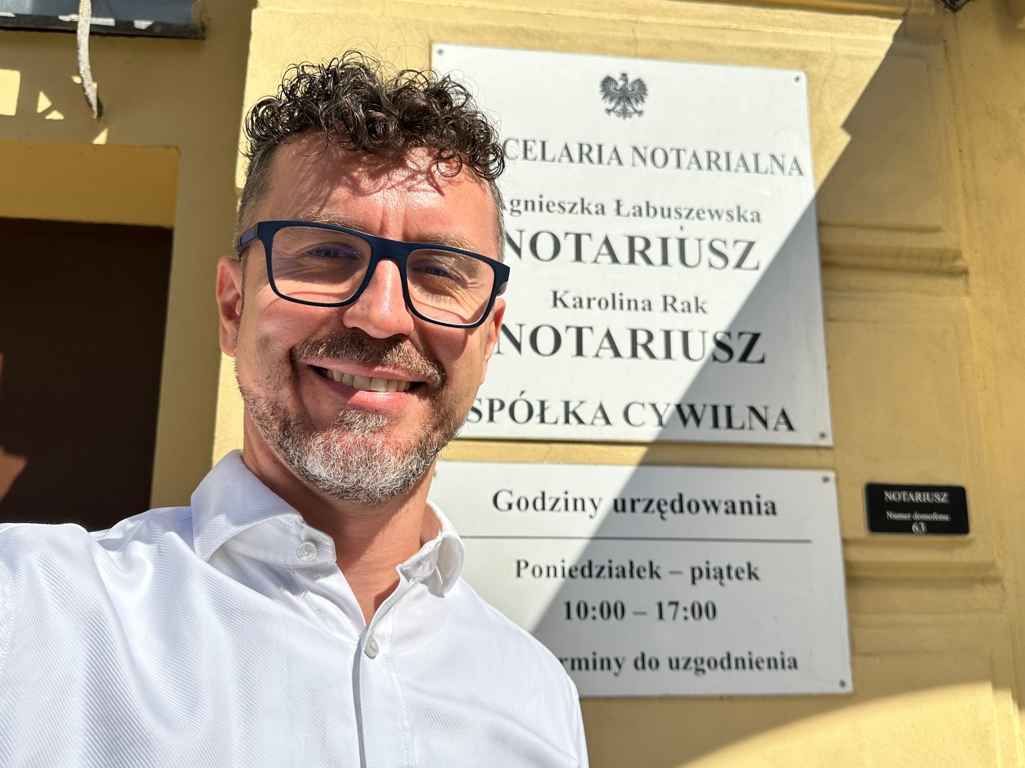 Uśmiechnięty mężczyzna w okularach robi selfie na tle tablicy informacyjnej kancelarii notarialnej Agnieszki Łabuszewskiej i Karoliny Rak. Tablica zawiera godziny urzędowania i numer domofonu.