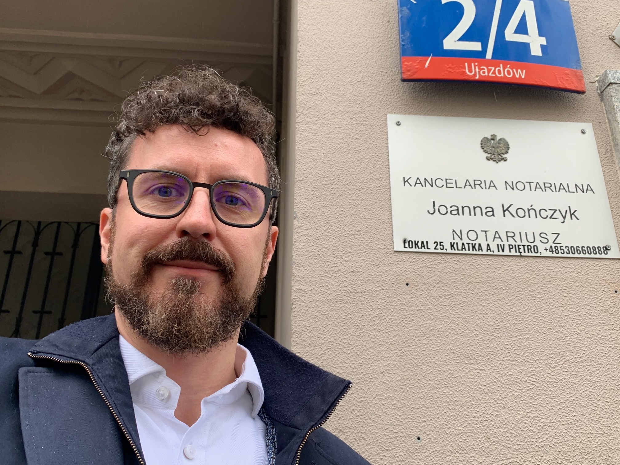 Portret mężczyzny w okularach na tle tabliczki z adresem Kancelarii Notarialnej Joanny Kończyk, z numerem budynku 2/4 przy ulicy Ujazdów.