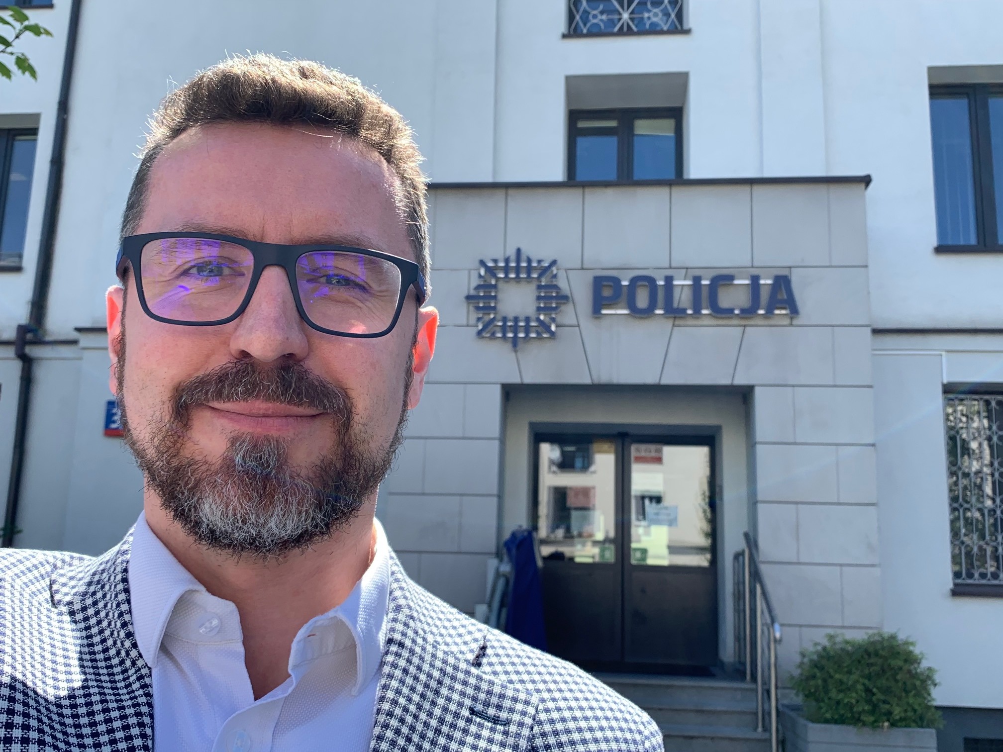Portret mężczyzny w okularach i marynarce w kratę na tle budynku z napisem POLICJA.