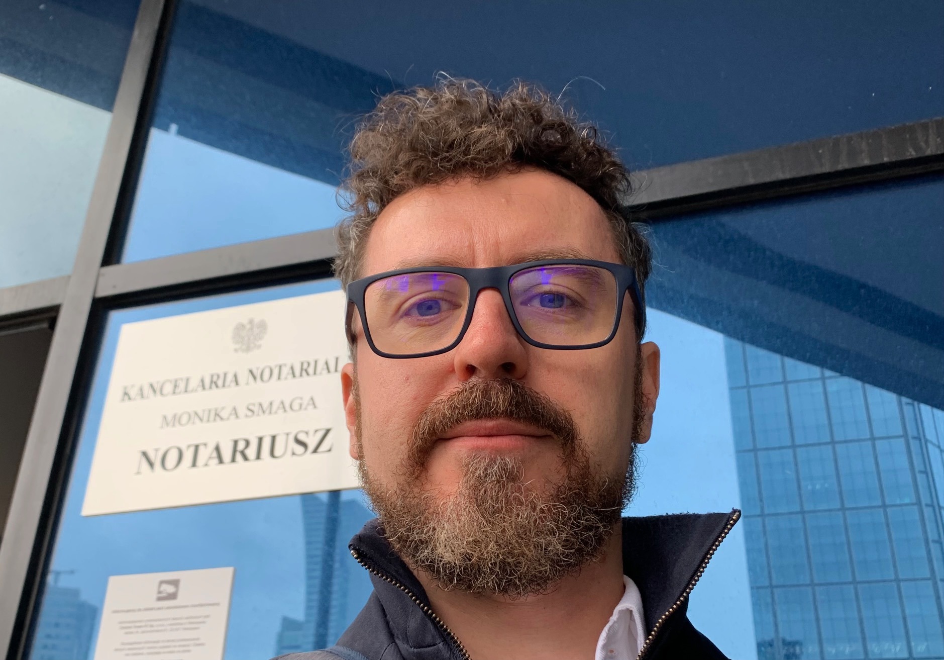 Autoportret mężczyzny w okularach na tle szyldu Kancelarii Notarialnej Moniki Smagi z widocznym budynkiem biurowym w odbiciu.