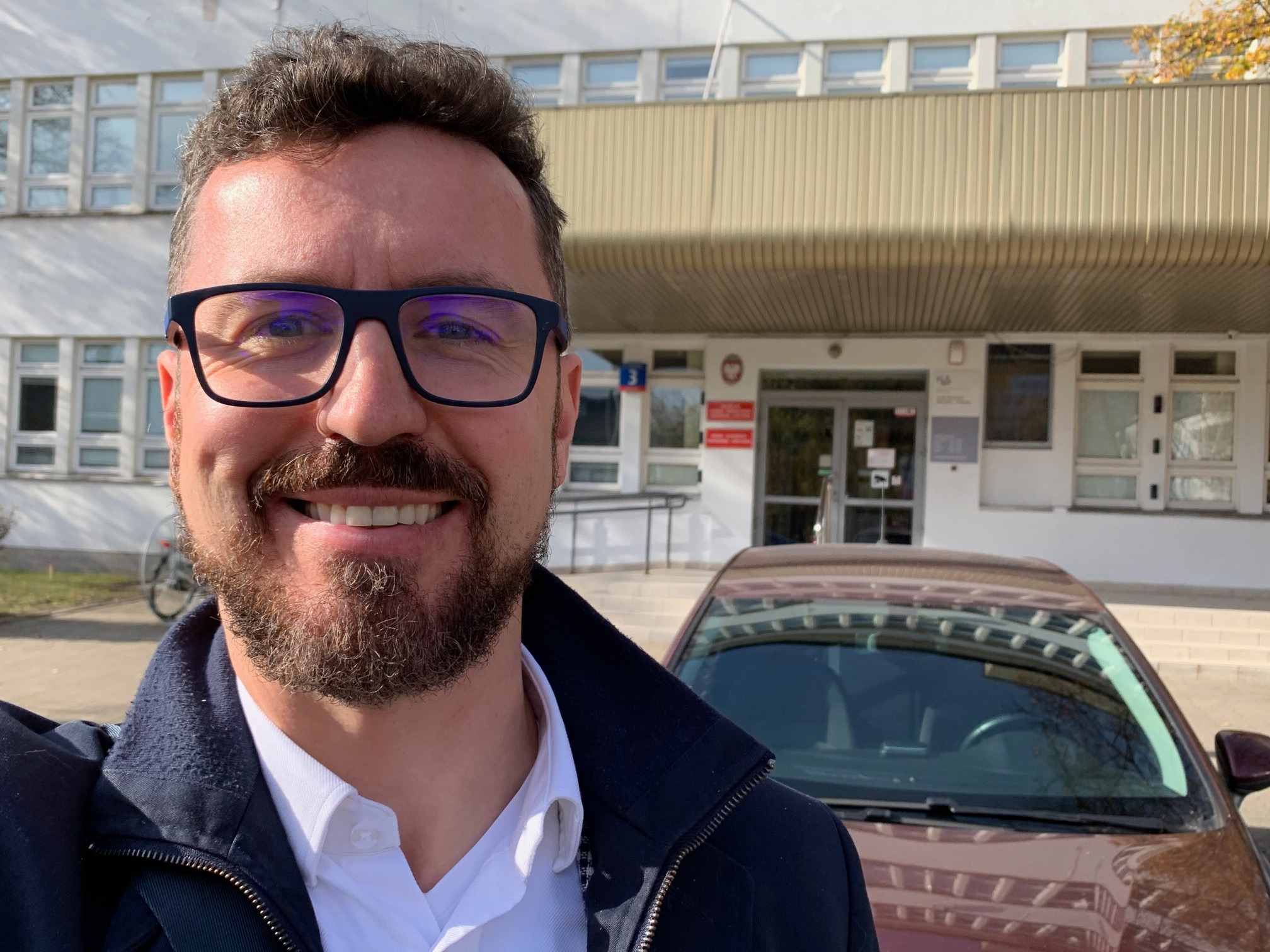 Uśmiechnięty mężczyzna w okularach robi selfie na tle budynku z tabliczkami i samochodu, prawdopodobnie przed urzędem lub sądem.