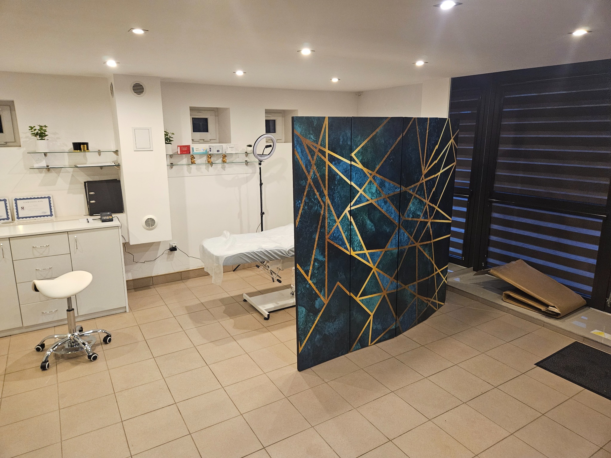 Wnętrze gabinetu z kozetką przykrytą jednorazowym prześcieradłem, białe meble i parawan z geometrycznym wzorem w odcieniach niebieskiego i złota.