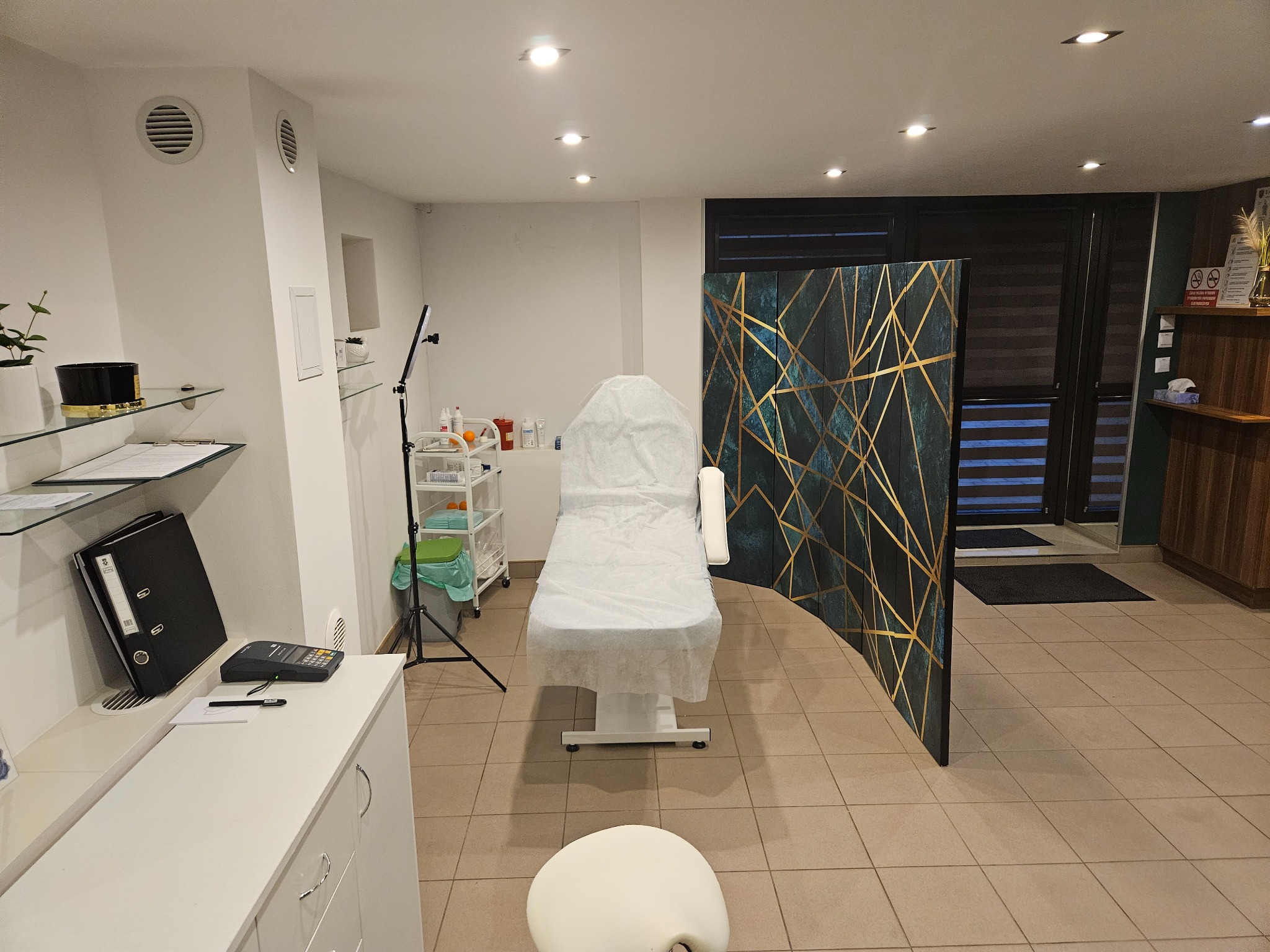 Jasne wnętrze gabinetu medycyny estetycznej z łóżkiem zabiegowym przykrytym białą narzutą, parawanem dekoracyjnym w kolorze ciemnej zieleni ze złotymi geometrycznymi wzorami oraz wyposażeniem...