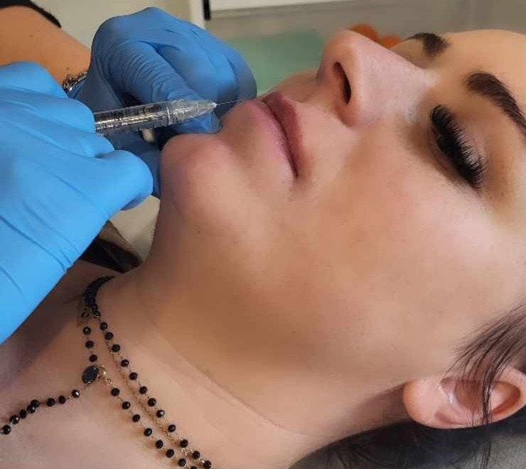 Iniekcja w usta pacjentki w rękawicach ochronnych, zbliżenie na twarz z widoczną igłą i kolczykiem z koralików.