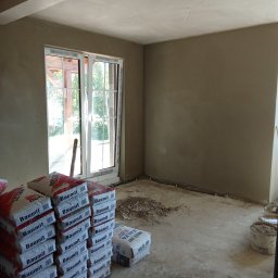 DOMPUR - Wnętrze po tynkowaniu, z oknem z widokiem na zieleń. Na podłodze worki z zaprawą Baumit i resztki materiałów budowlanych. Ściany gładkie, przygotowane do malowania.