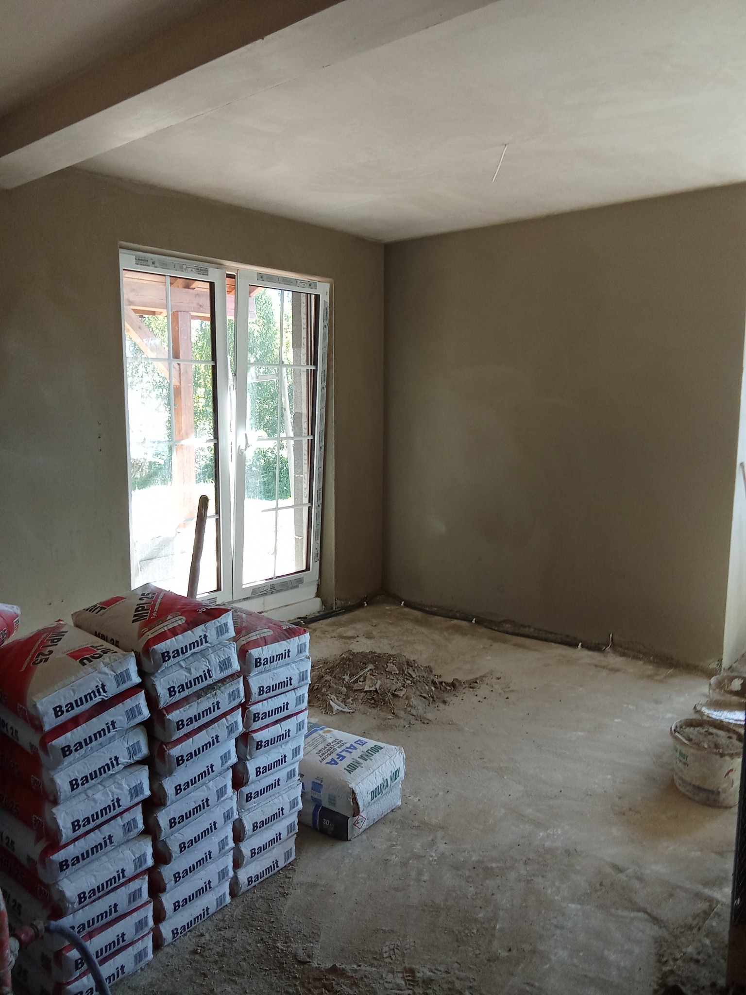 Wnętrze po tynkowaniu, z oknem z widokiem na zieleń. Na podłodze worki z zaprawą Baumit i resztki materiałów budowlanych. Ściany gładkie, przygotowane do malowania.