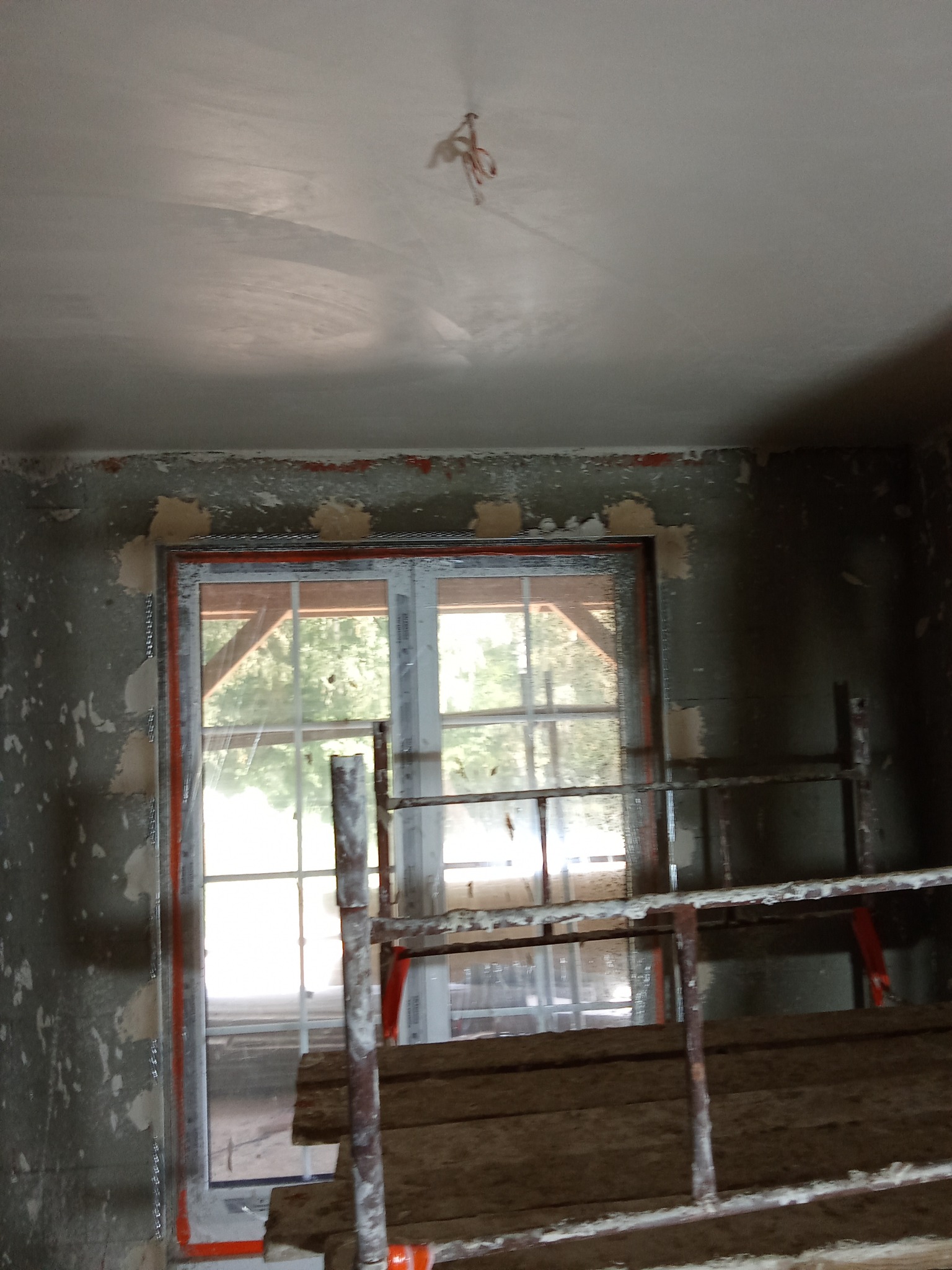 Wnętrze w trakcie ocieplania pianką PUR: ściany z resztkami starej farby, okno z widoczną konstrukcją dachu, rusztowanie i świeżo nałożona pianka na suficie.