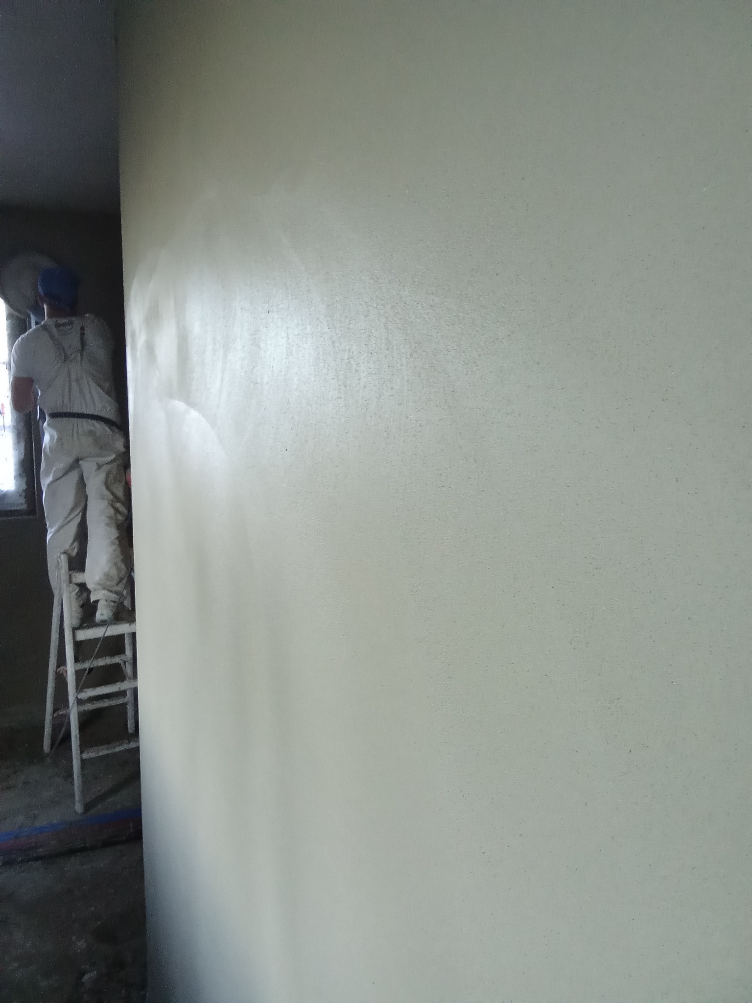 Świeżo nałożona warstwa izolacji na ścianie wewnętrznej, pracownik na drabinie w tle, widoczne ślady aplikacji natryskowej.