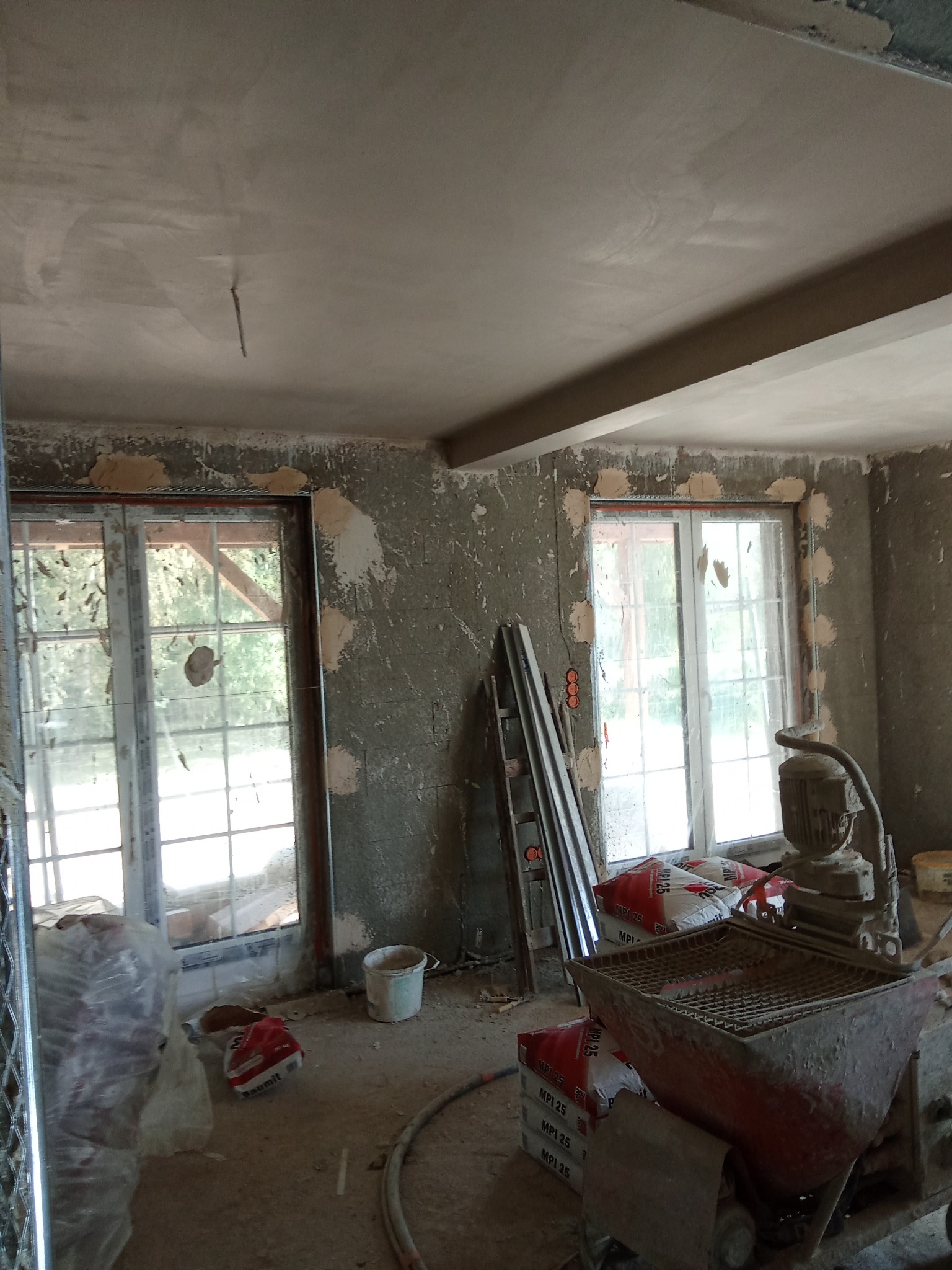 Wnętrze w trakcie prac wykończeniowych: ściany z naniesioną zaprawą, worki z materiałami budowlanymi, maszyna do tynkowania i drabina oparta o ścianę.