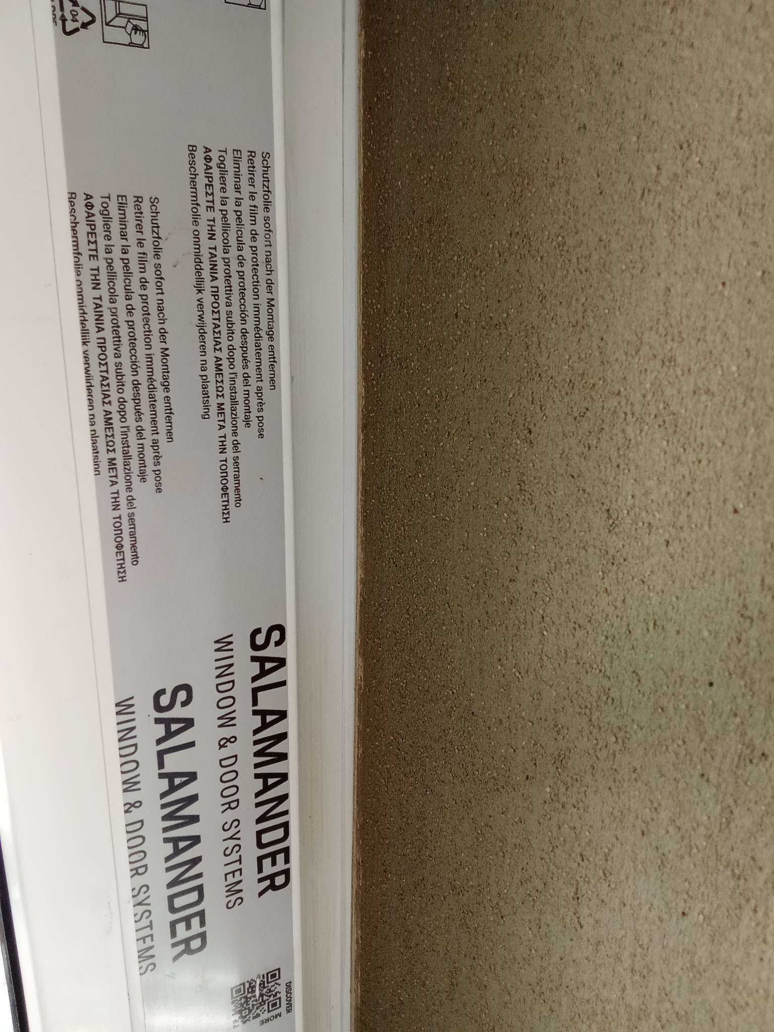 Fragment okna Salamander z instrukcją usunięcia folii ochronnej po montażu, na tle ściany o szorstkiej fakturze. Widoczny kod QR.