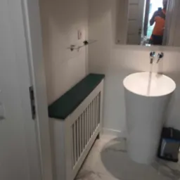 Biała, wolnostojąca umywalka cylindryczna z chromowaną armaturą w łazience z marmurową podłogą, obok zabudowa grzejnika z zielonym blatem i szklana półka na ścianie, widoczne odbicie osoby w lustrze.