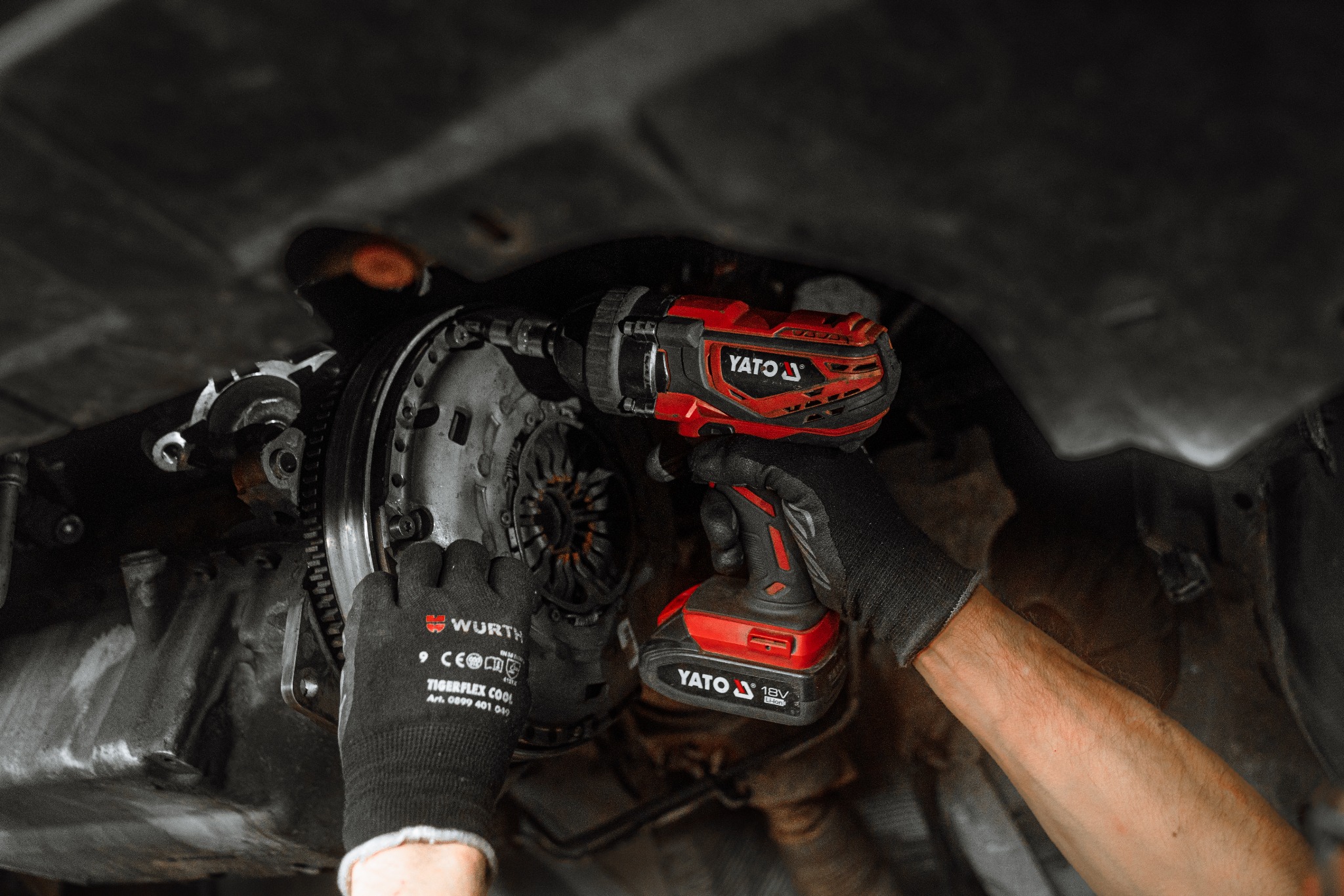 Czarno-czerwony akumulatorowy klucz udarowy Yato w dłoniach mechanika w rękawicach Wurth, podczas pracy przy sprzęgle samochodu w Ostrowcu Świętokrzyskim.