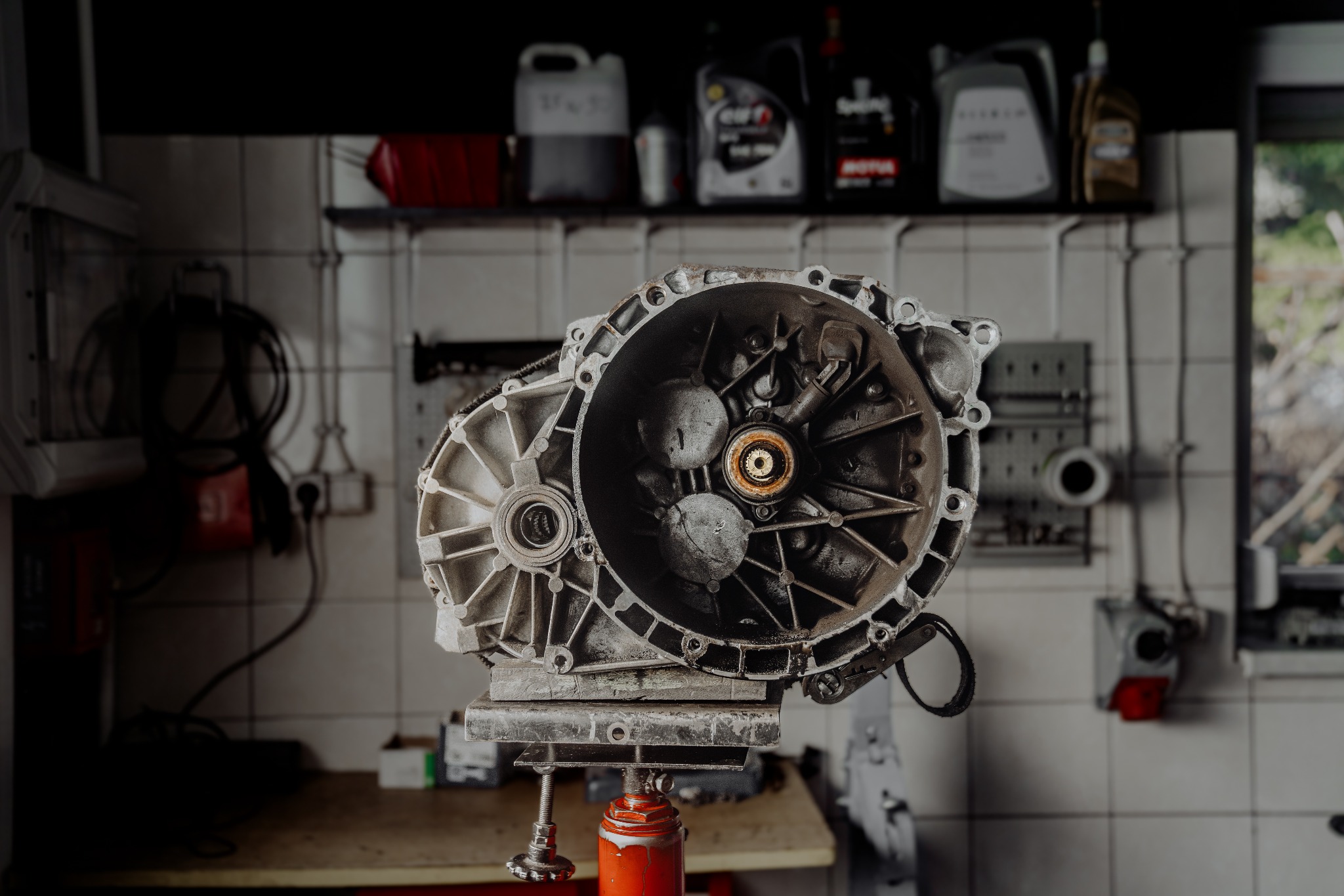 Zdjęcie skrzyni biegów samochodowej, zamontowanej na czerwonym podnośniku warsztatowym, widoczne oleje silnikowe na półce w tle, warsztat z białymi kafelkami.