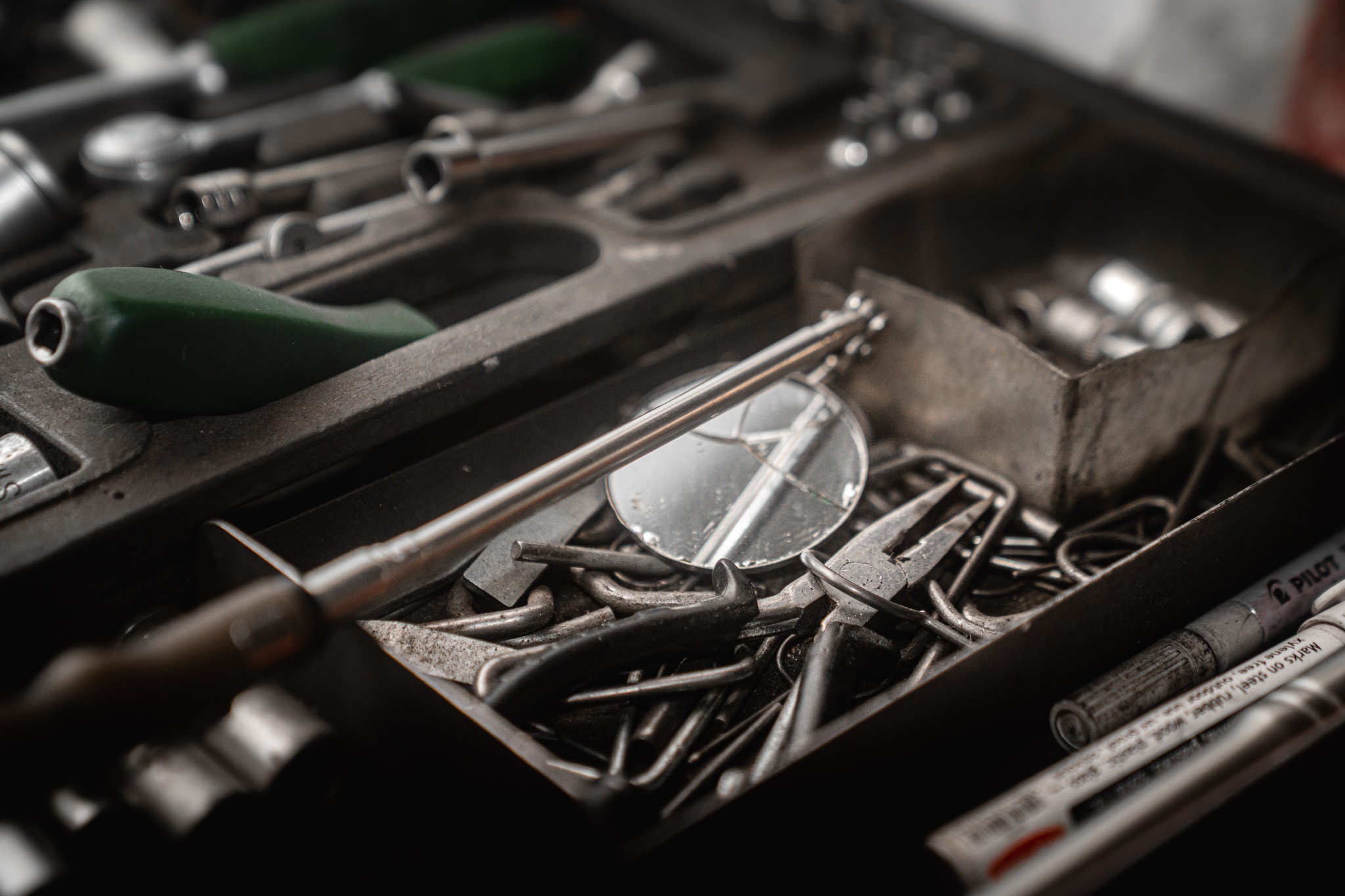 Otwarty organizer warsztatowy z różnorodnymi narzędziami, w tym klucze nasadowe, śrubokręt z zieloną rękojeścią, lusterko inspekcyjne, szczypce i markery. Narzędzia są używane i noszą ślady...