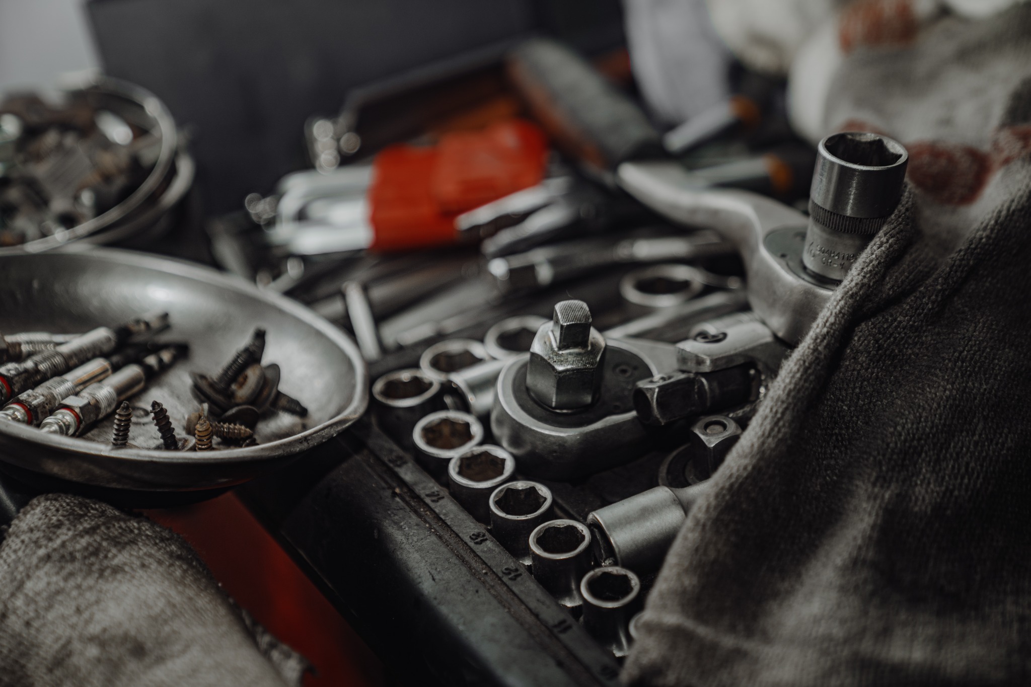 Zestaw kluczy nasadowych, grzechotka, metalowa miseczka z wkrętami i świecami zapłonowymi na warsztatowym stole, przykryty szarą szmatką.