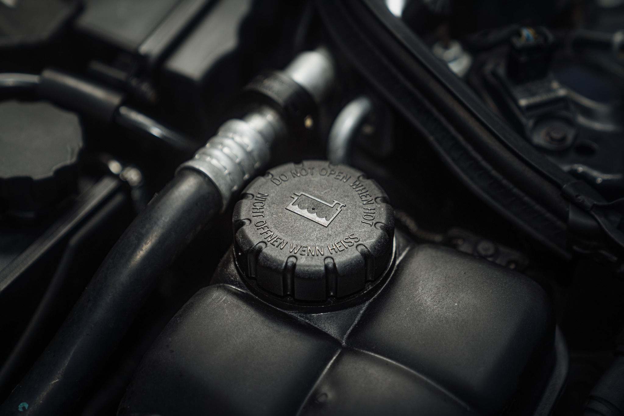 Zbliżenie na zbiornik wyrównawczy płynu chłodniczego w samochodzie z napisem ostrzegawczym 'Do not open when hot' w języku angielskim i niemieckim.