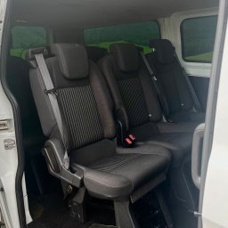 Paskal Zgierski F.H.U „EasyBUS” - Wnętrze białego busa z trzema rzędami czarnych foteli z pasami bezpieczeństwa, widok z otwartych drzwi bocznych na zielony krajobraz za oknem.