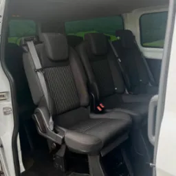 Wnętrze białego busa z trzema rzędami czarnych foteli z pasami bezpieczeństwa, widok z otwartych drzwi bocznych na zielony krajobraz za oknem.