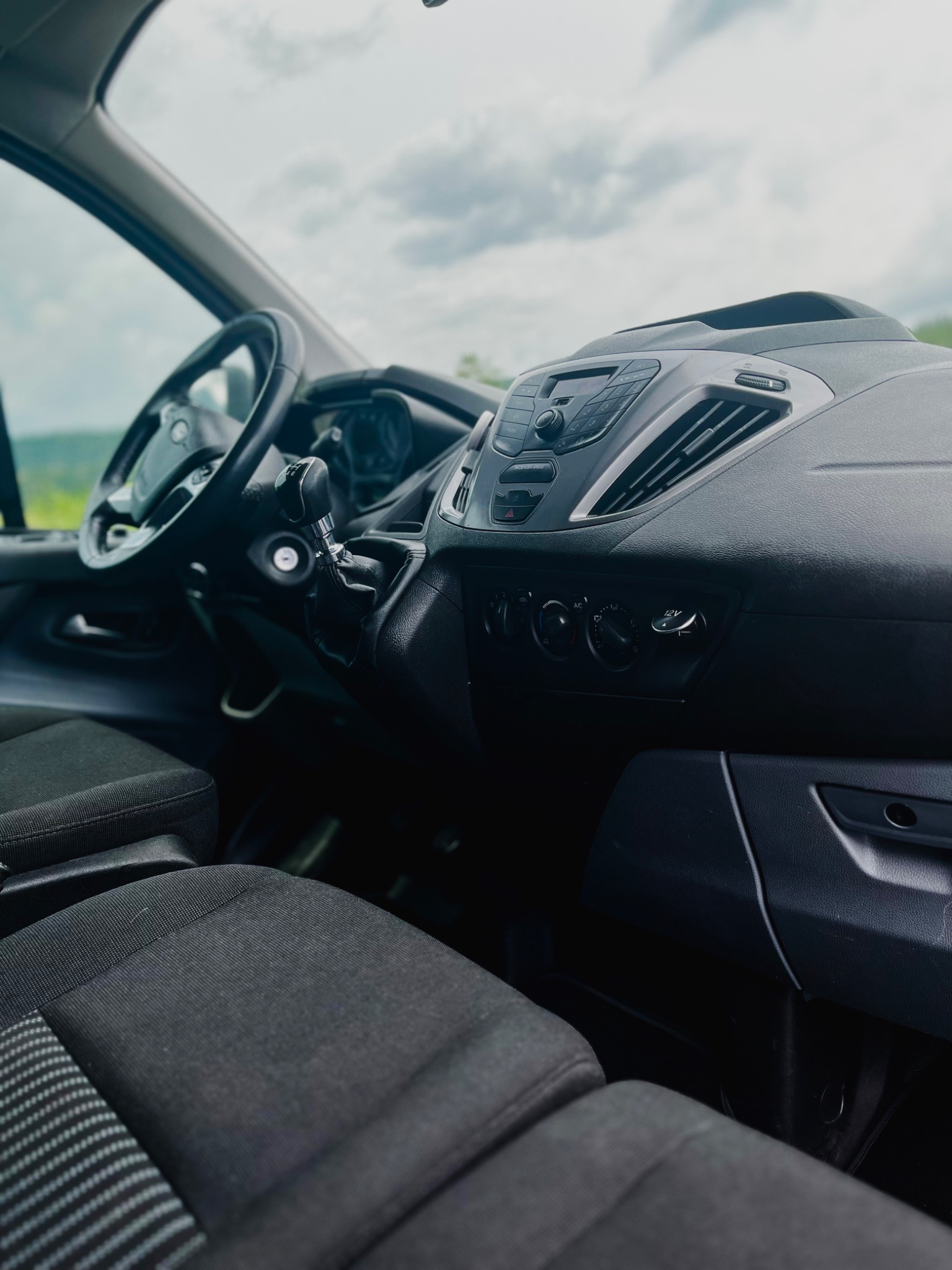 Wnętrze kabiny busa: deska rozdzielcza, kierownica i fotel kierowcy, widok z perspektywy pasażera, w tle pochmurne niebo.